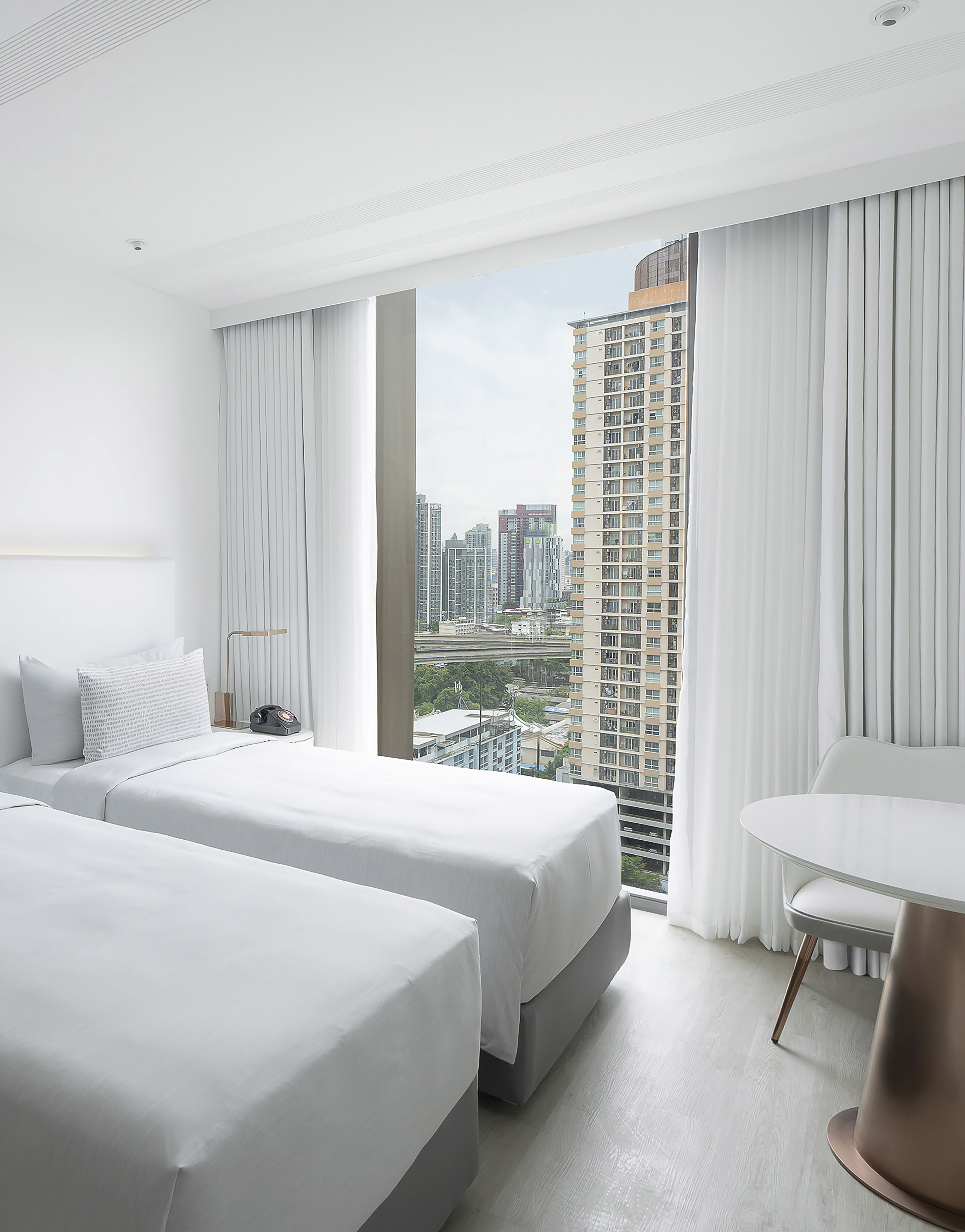 a room with two beds and a table and a window with tall buildings in the background