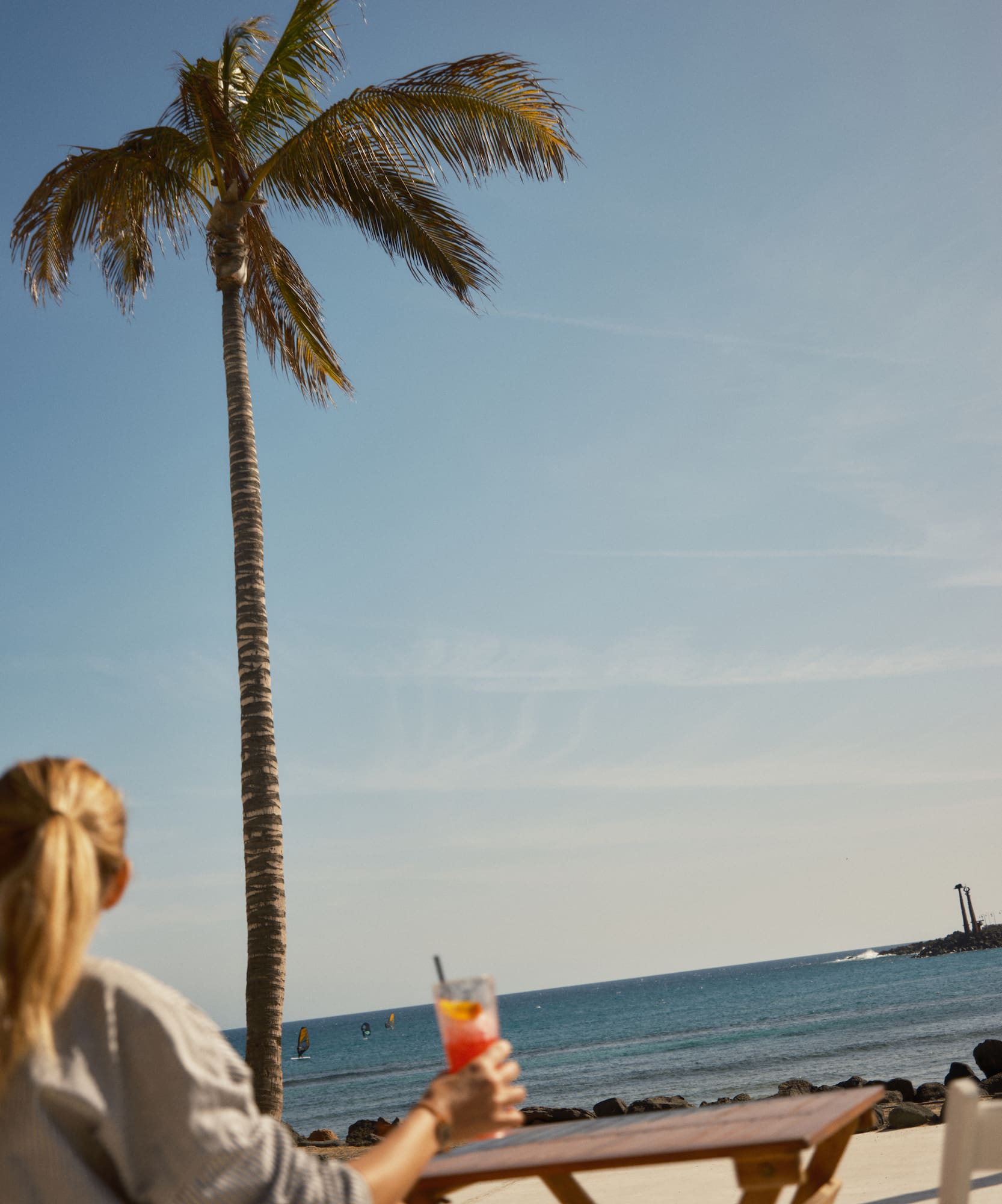 Blonde woman holding cocktail, watching windsurfers on tropical ocean by a palm tree.