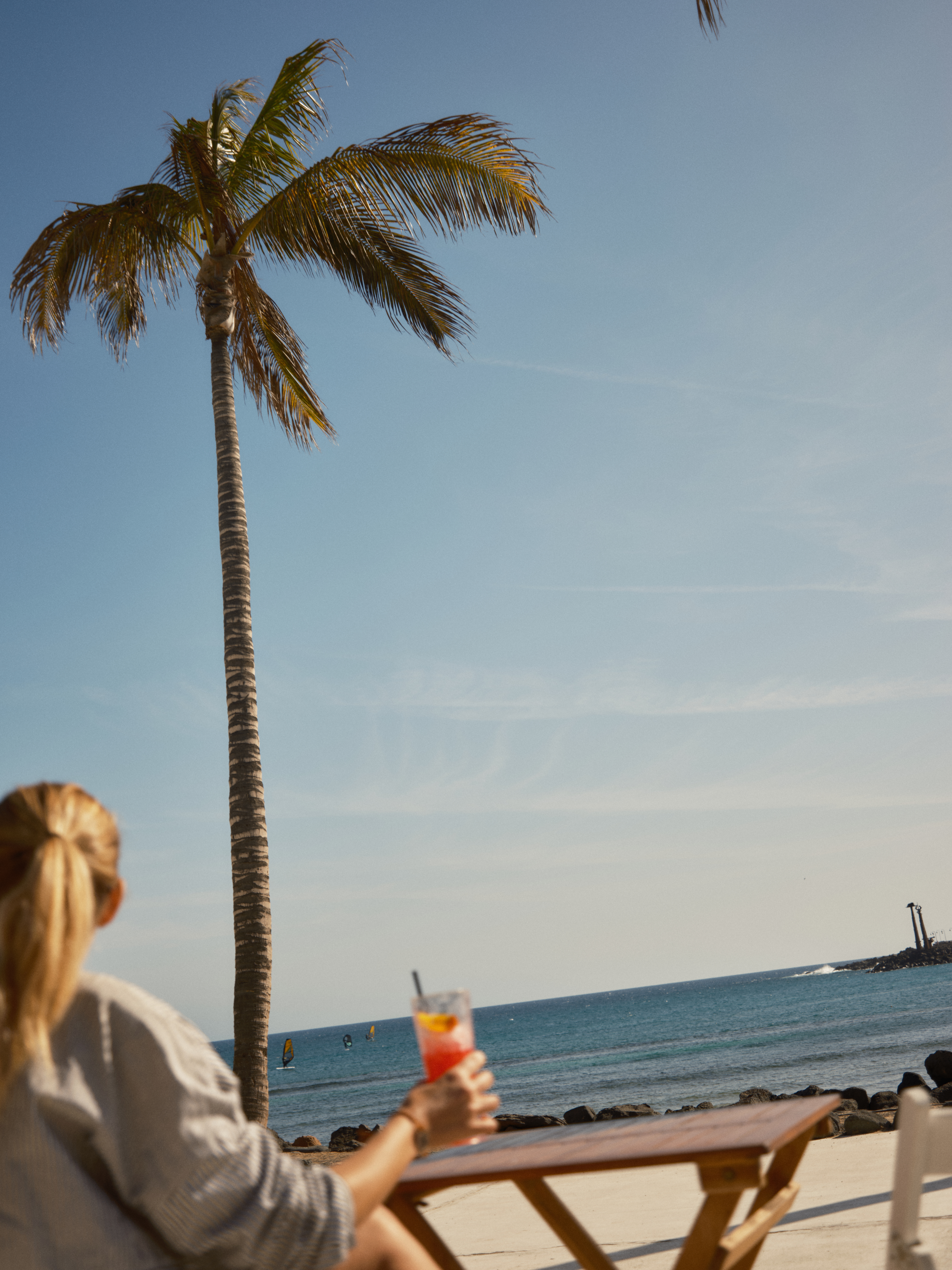 Blonde woman holding cocktail, watching windsurfers on tropical ocean by a palm tree.