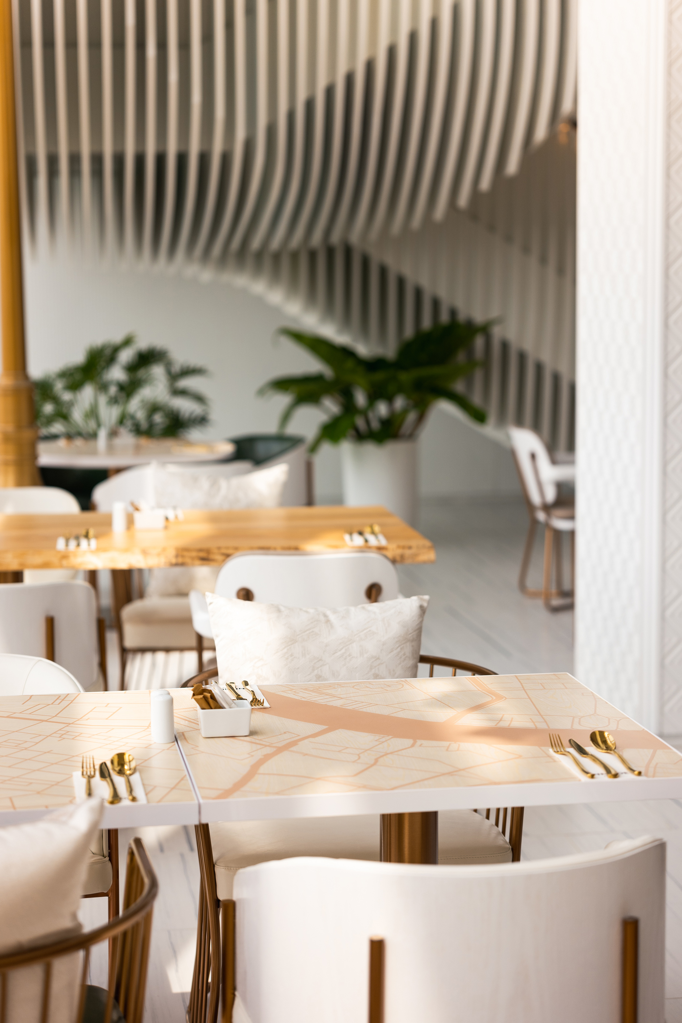 Modern restaurant interior: map tables, white chairs, gold cutlery, slatted ceiling.