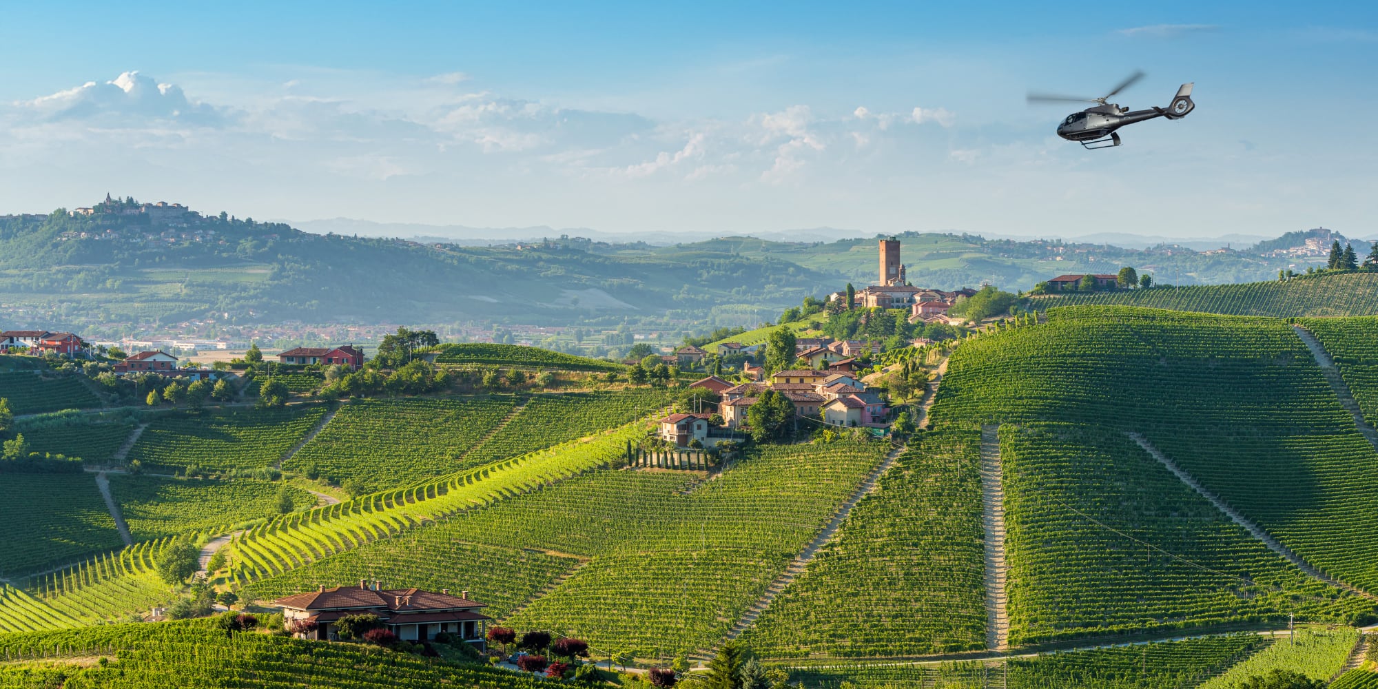 Helicopter over rolling green vineyards and hilltop village in a sunny wine region.