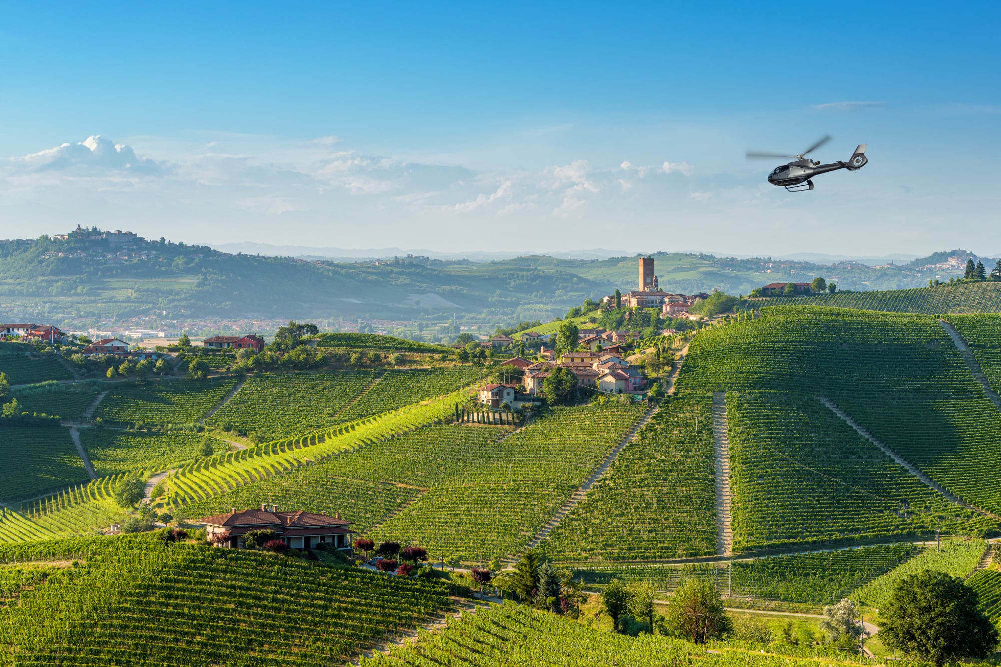 Helicopter over rolling green vineyards and hilltop village in a sunny wine region.