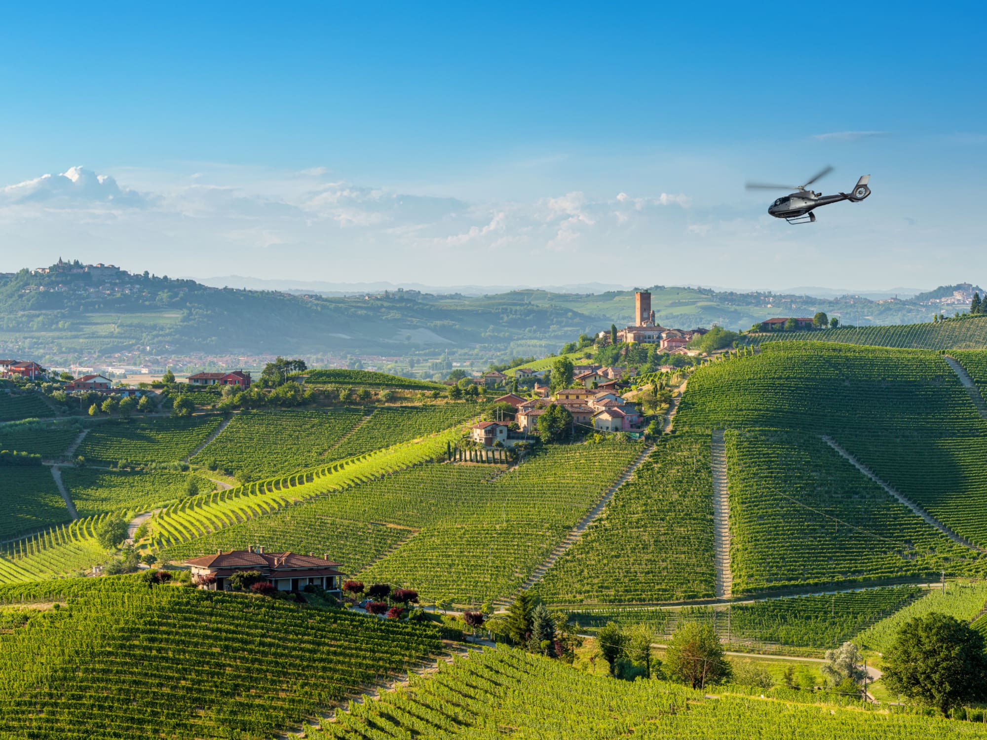 Helicopter over rolling green vineyards and hilltop village in a sunny wine region.