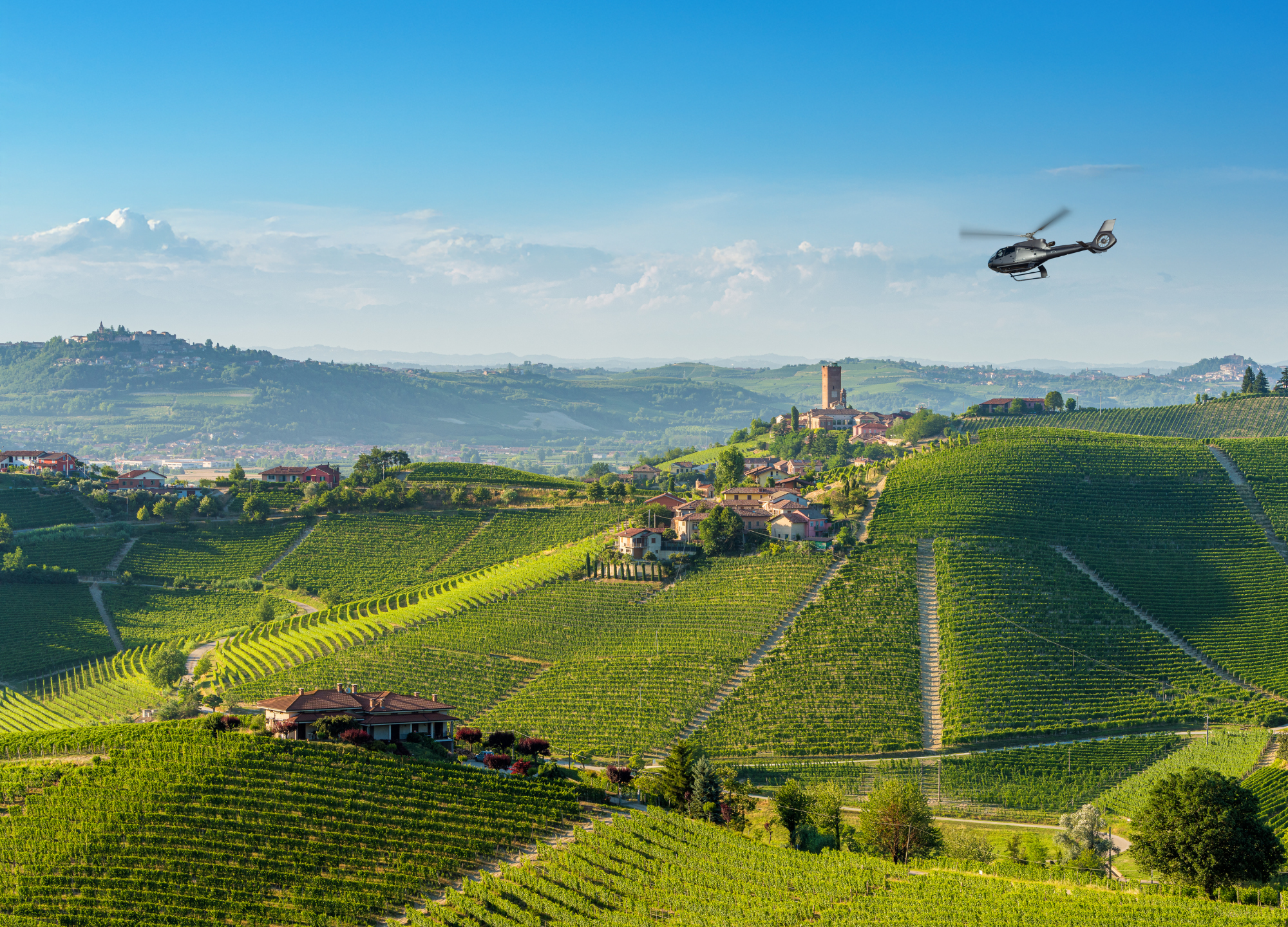 Helicopter over rolling green vineyards and hilltop village in a sunny wine region.