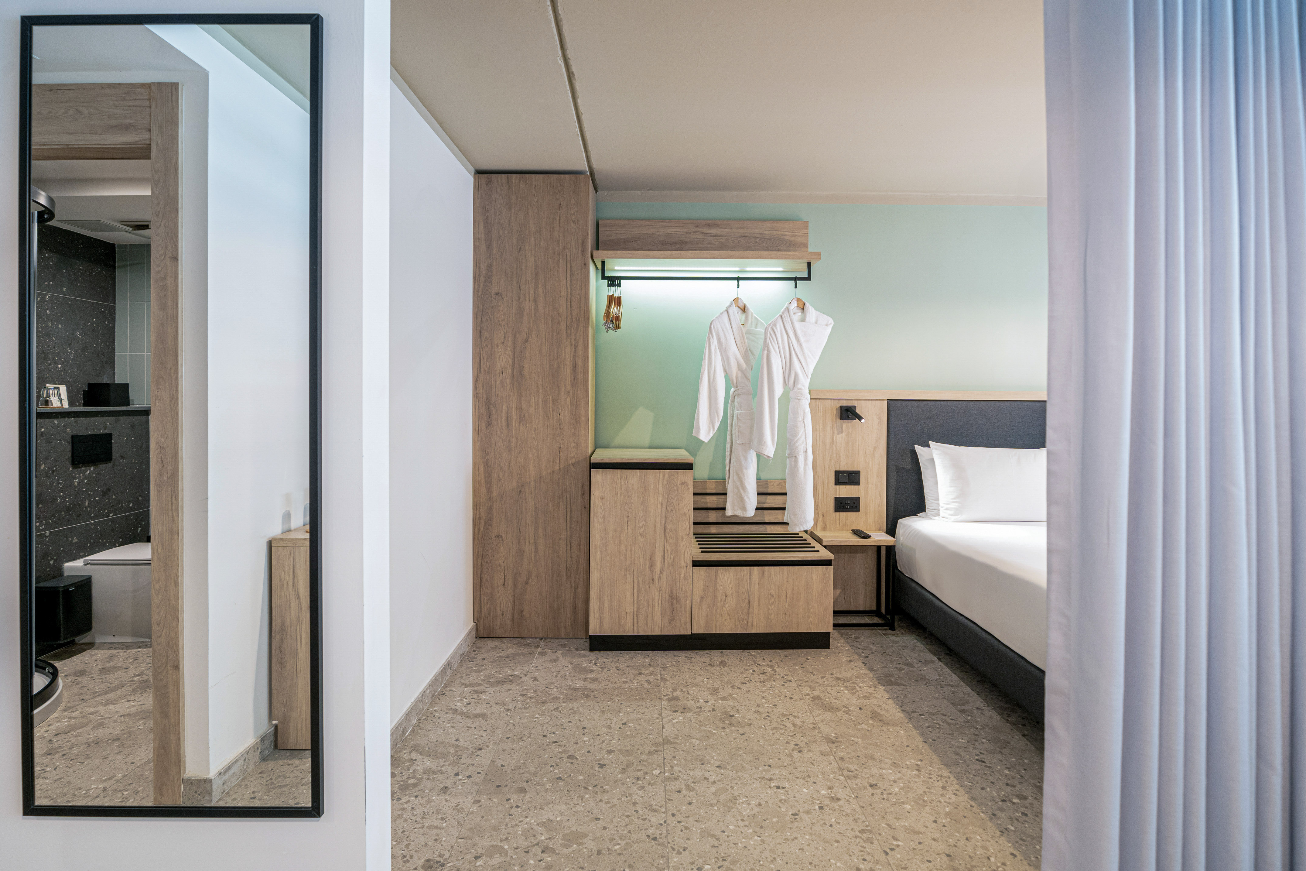 Minimalist hotel room with bed, wooden wardrobe, bathrobes, and a mirrored bathroom.