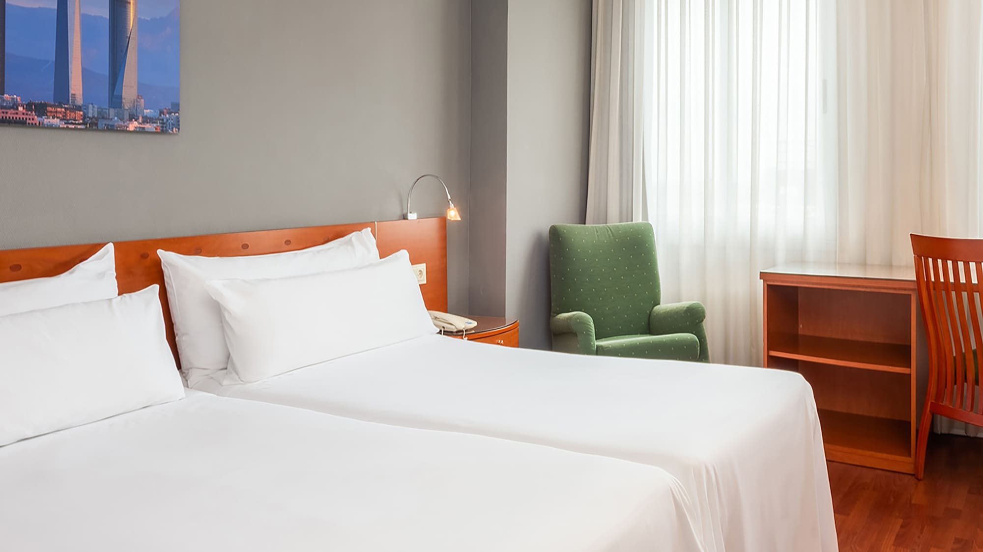 Cozy hotel room with twin beds, green armchair, desk, and cityscape art.