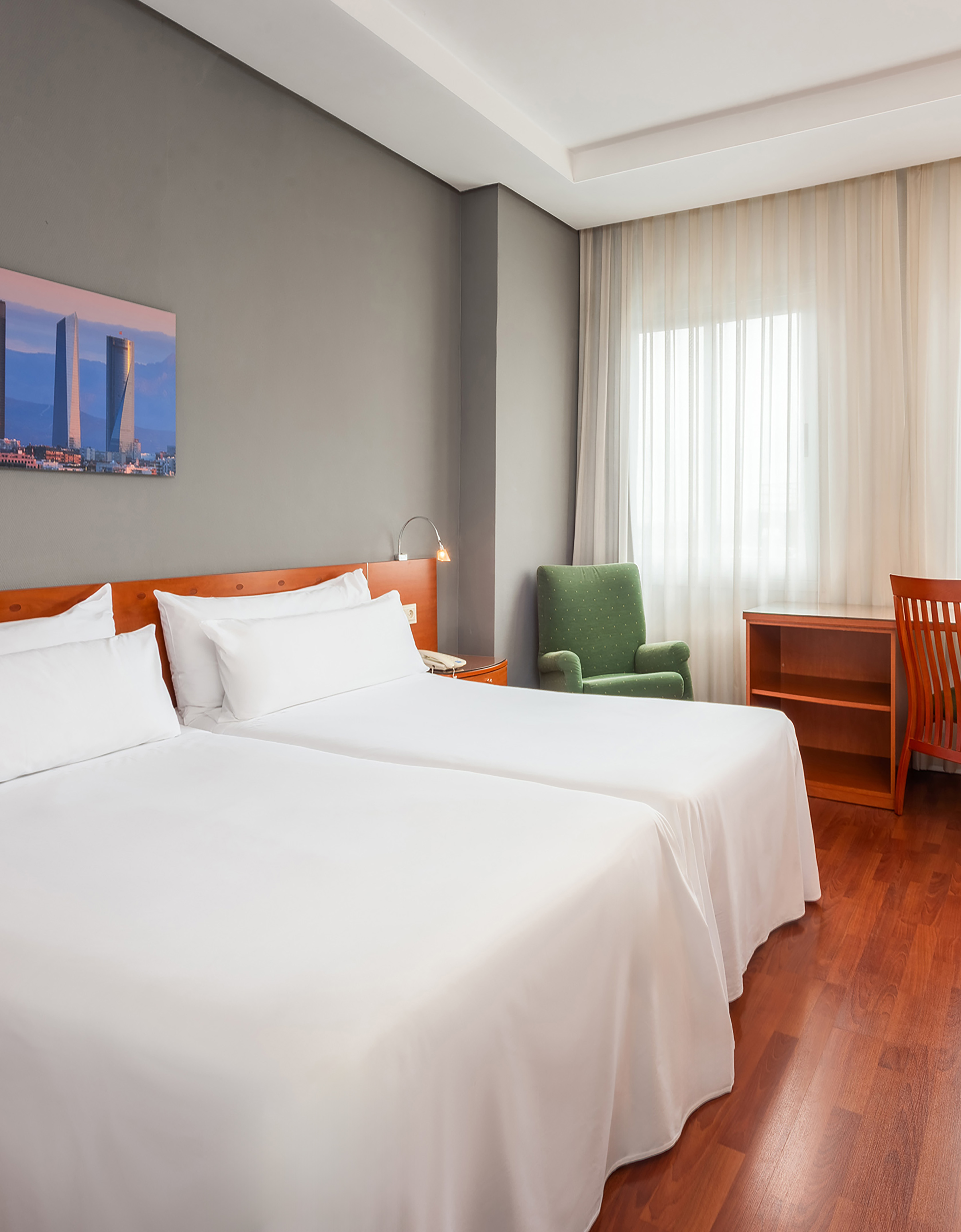 Cozy hotel room with twin beds, green armchair, desk, and cityscape art.