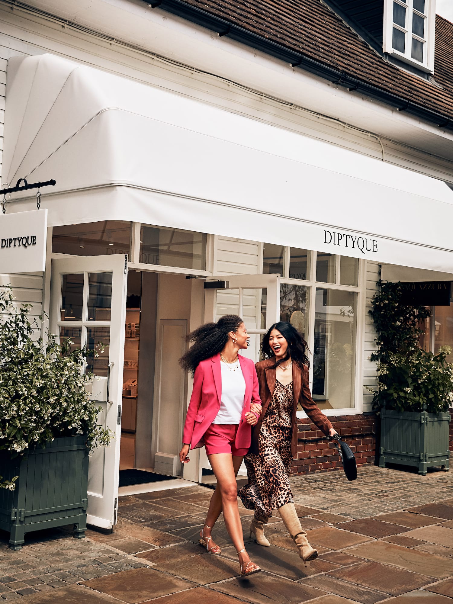 Two stylish women walk smiling past Diptyque boutique entrance.