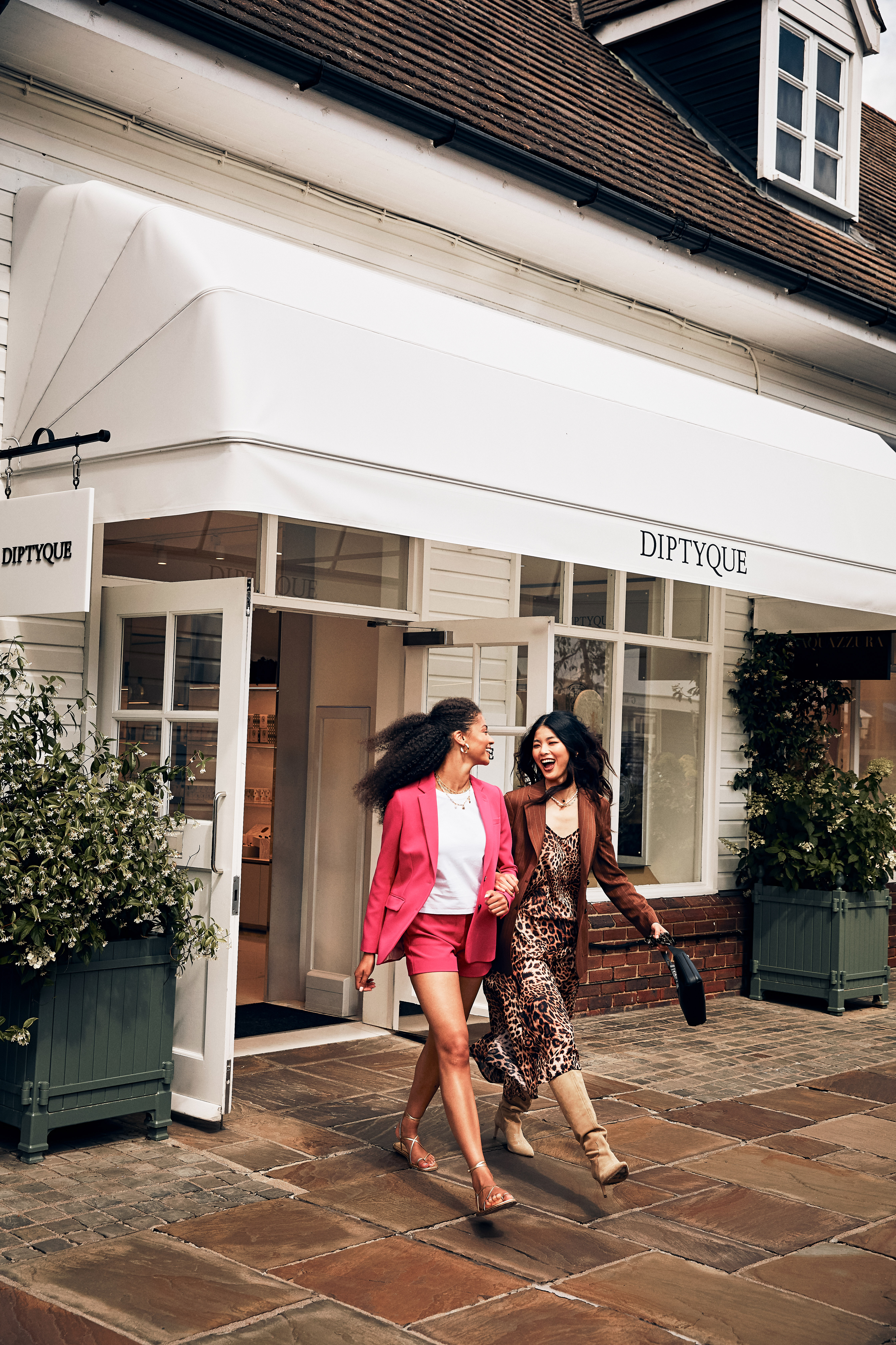 Two stylish women walk smiling past Diptyque boutique entrance.
