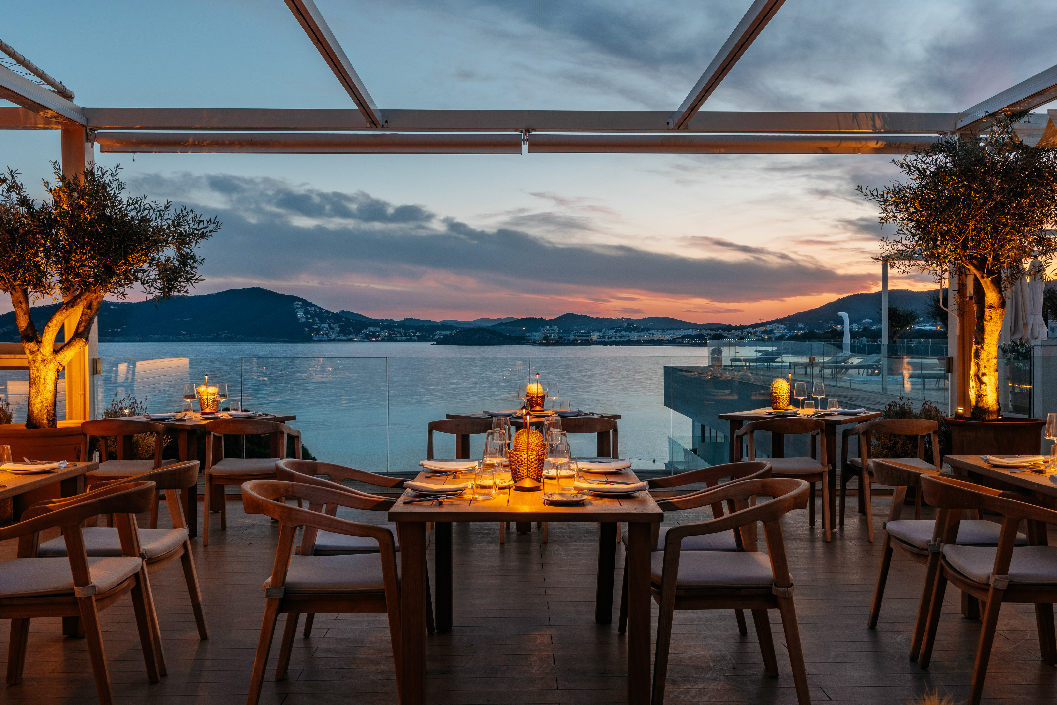 Rooftop restaurant with ocean view at sunset, set tables, and olive trees.