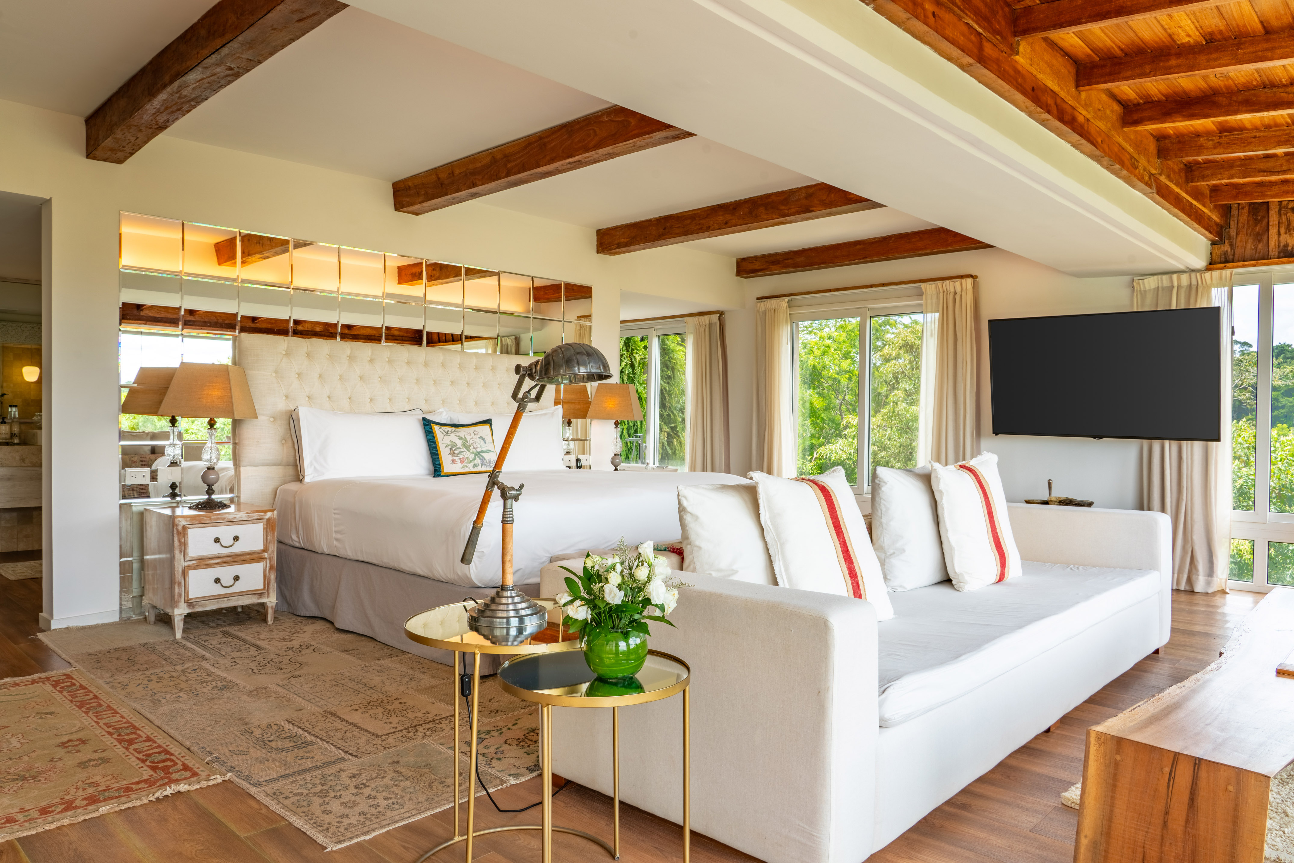 Luxury bedroom with king bed, exposed wood beams, sofa, and lush nature view.