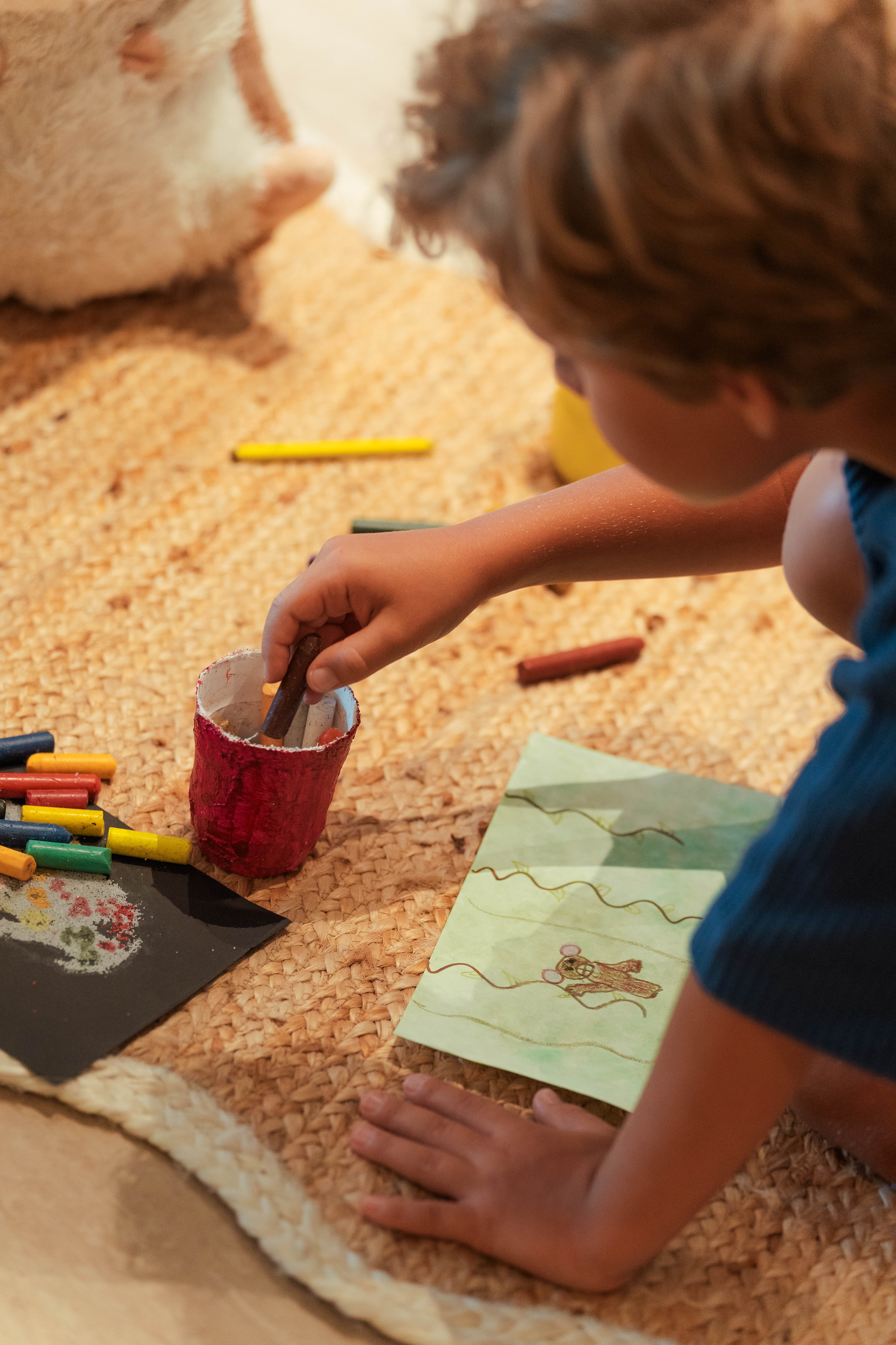 Niño coloreando con crayones en papel sobre alfombra yute. Actividad infantil creativa.