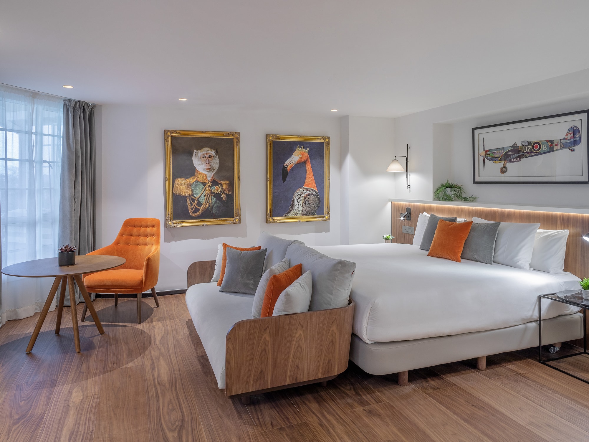 Modern hotel room with large bed, sofa, orange armchair, and eclectic artwork.