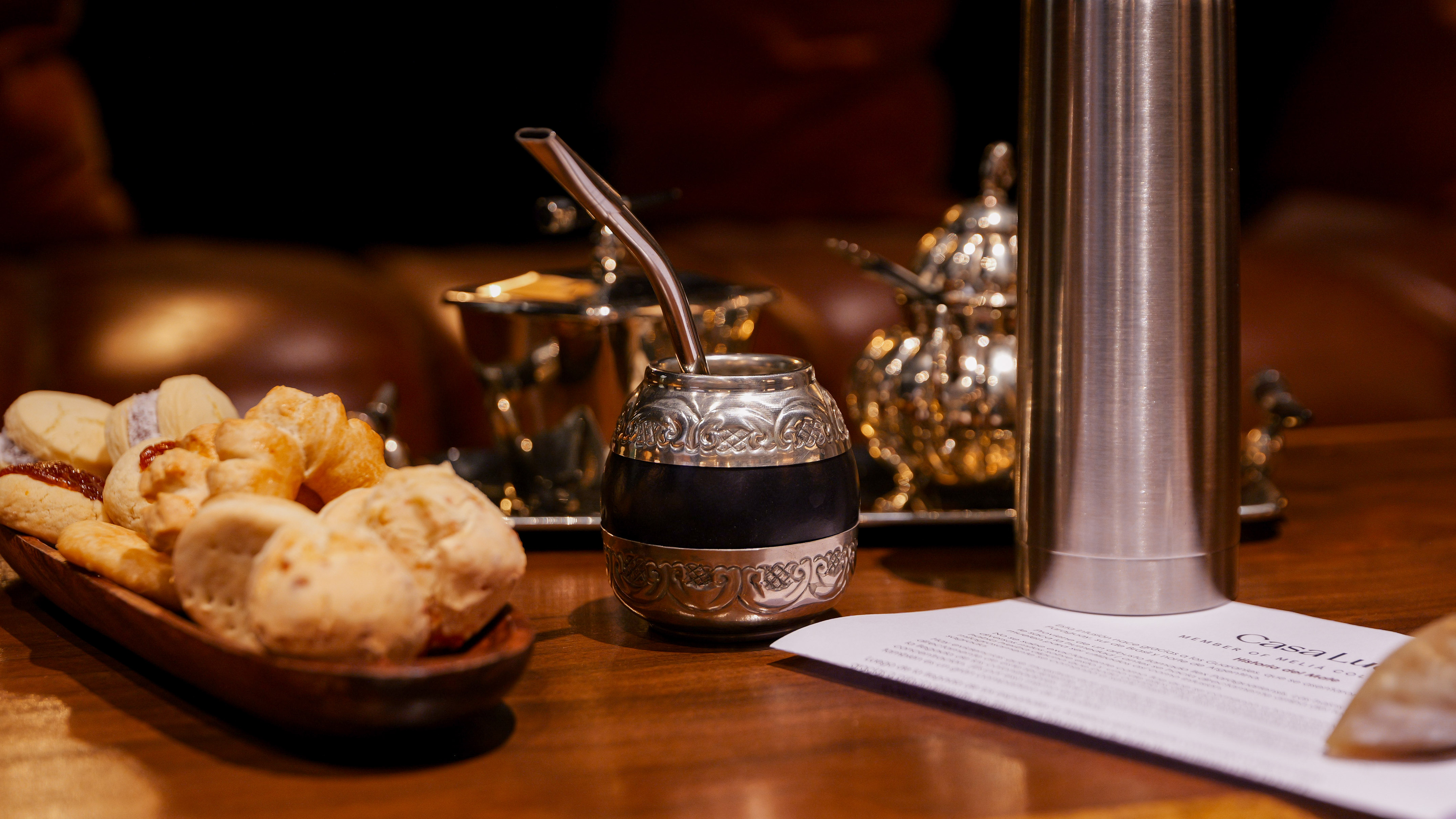 Silver mate gourd with bombilla, thermos, and South American pastries on a table.