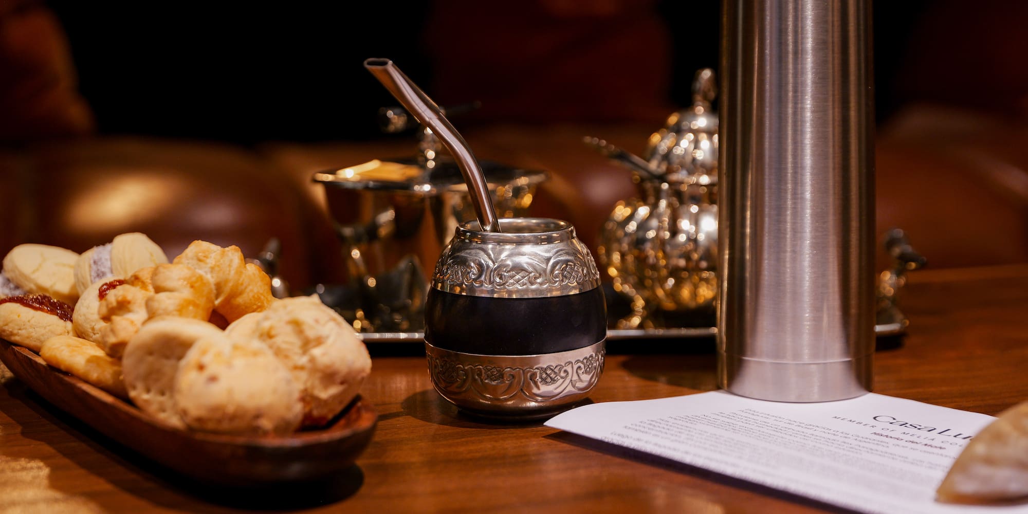 Silver mate gourd with bombilla, thermos, and South American pastries on a table.