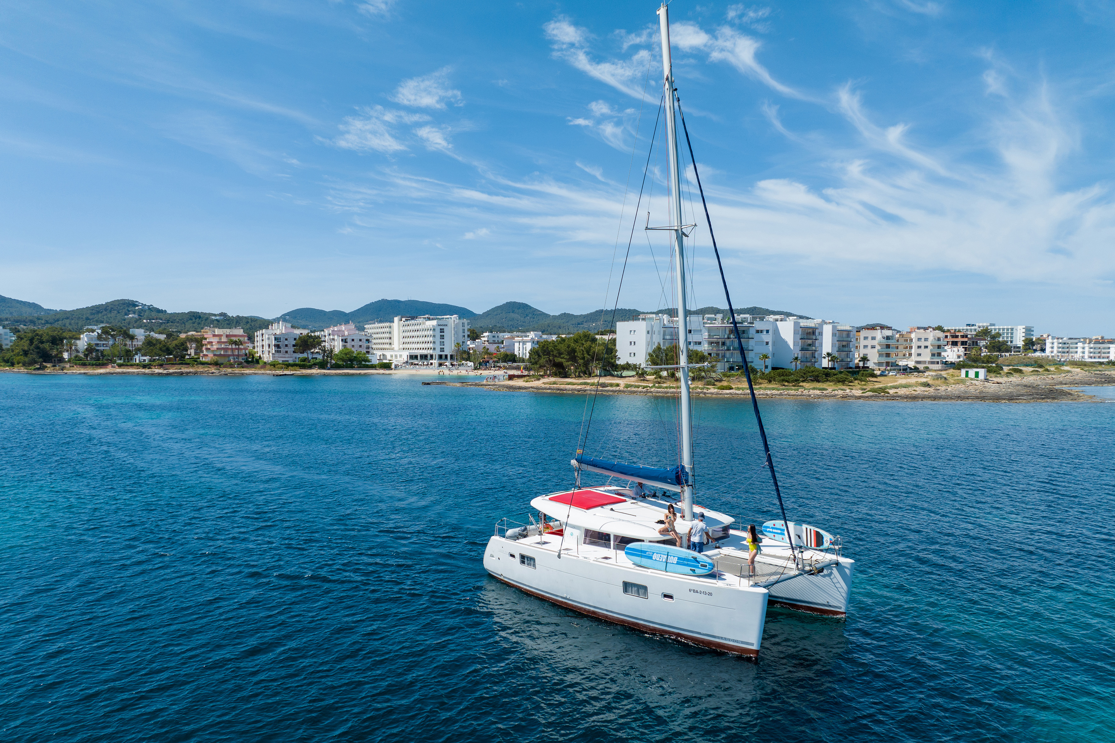 White catamaran yacht on clear blue sea, coastal resort in background.