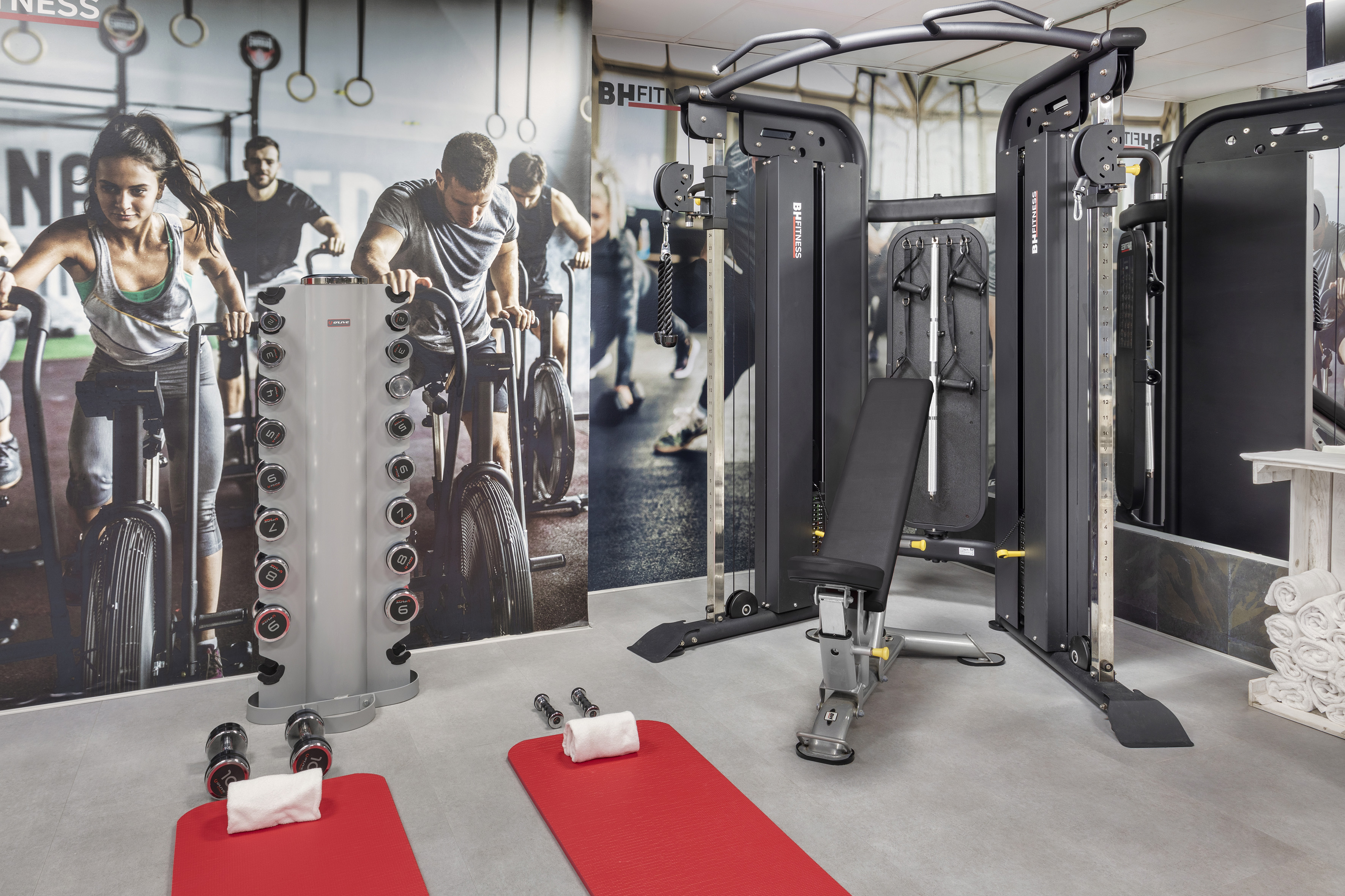 Modern gym with BH FITNESS cable machine, weight bench, dumbbell rack, and red exercise mats.