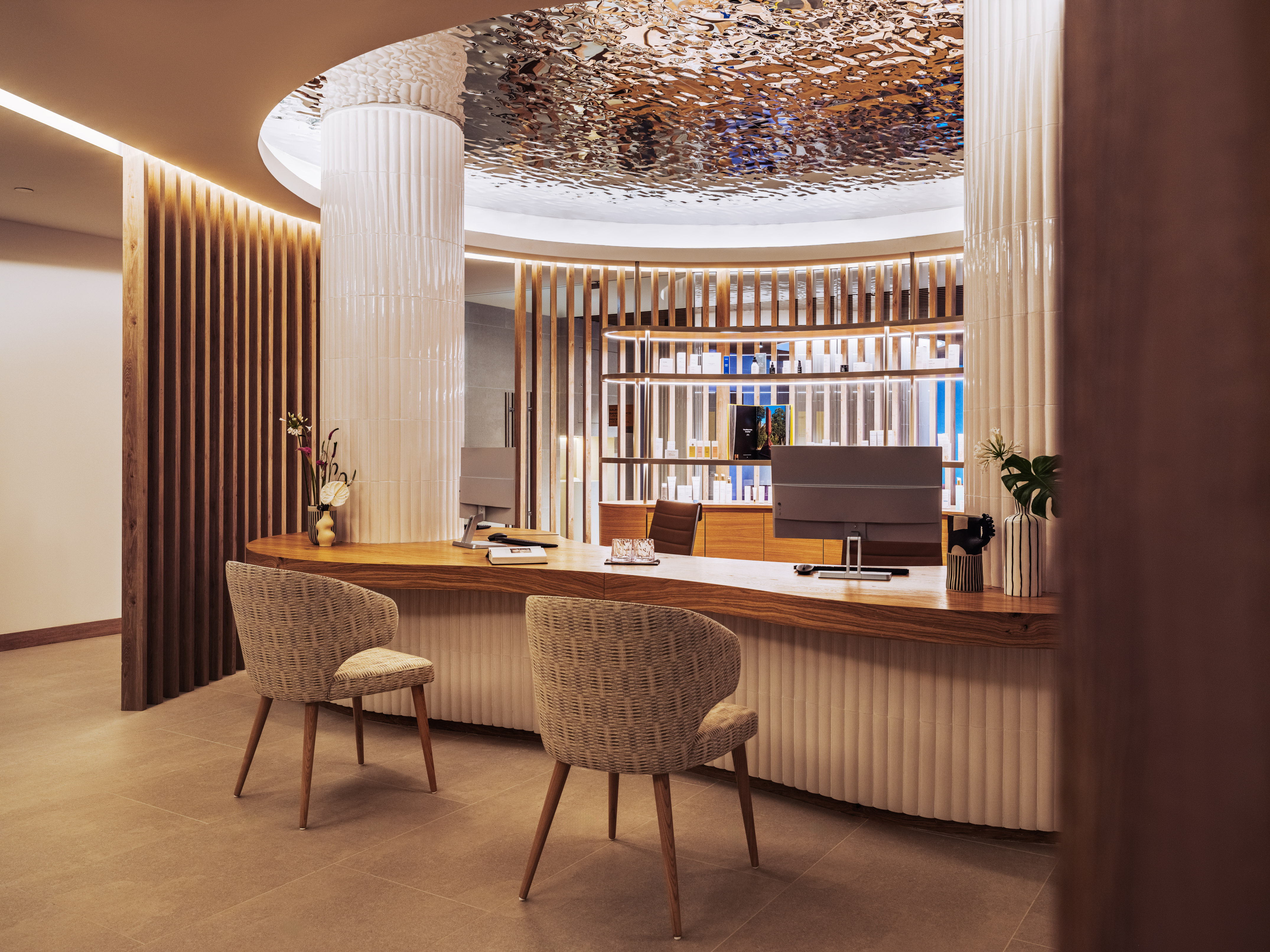 Modern spa reception, curved wood desk, woven chairs, reflective ceiling.
