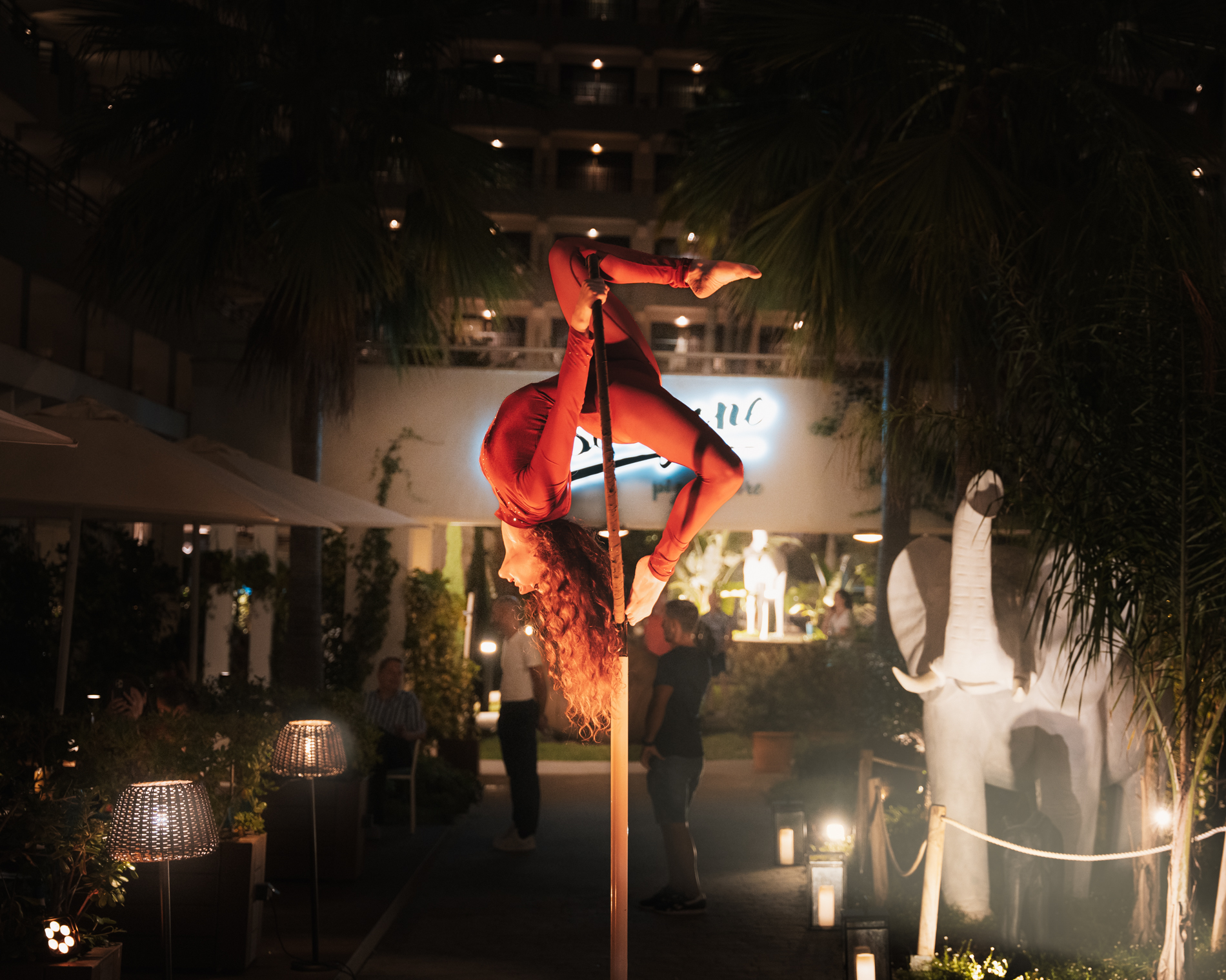 Aerialist in red performs pole acrobatics at night event with palm trees.