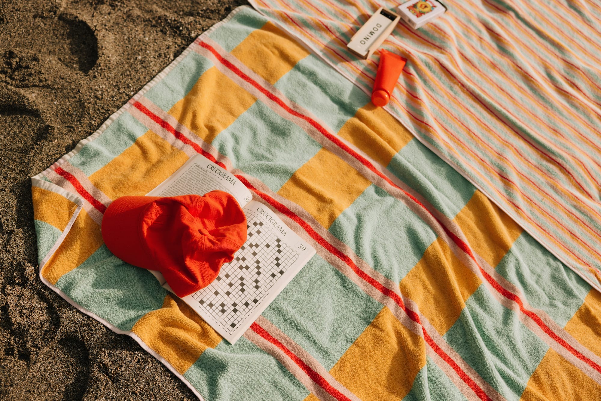 Colorful striped beach towel with red cap, crossword, sunscreen, dominoes on sand.