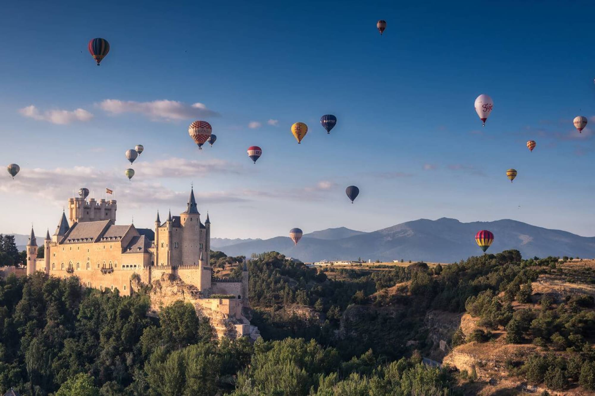 A castle sits atop a hill, surrounded by a sky full of hot air balloons.