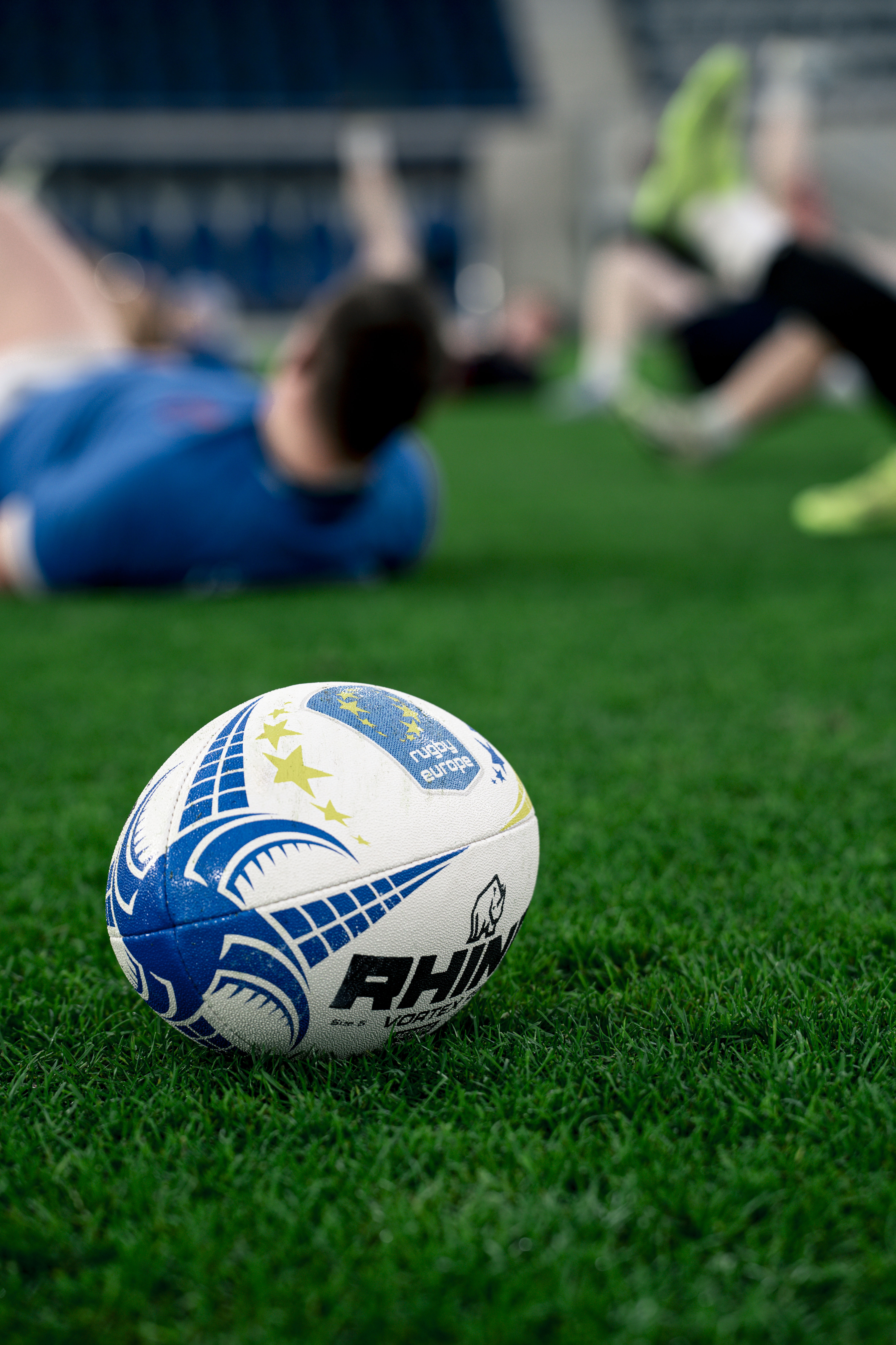 Rhino Rugby Europe ball on green grass, blurred players resting in background.