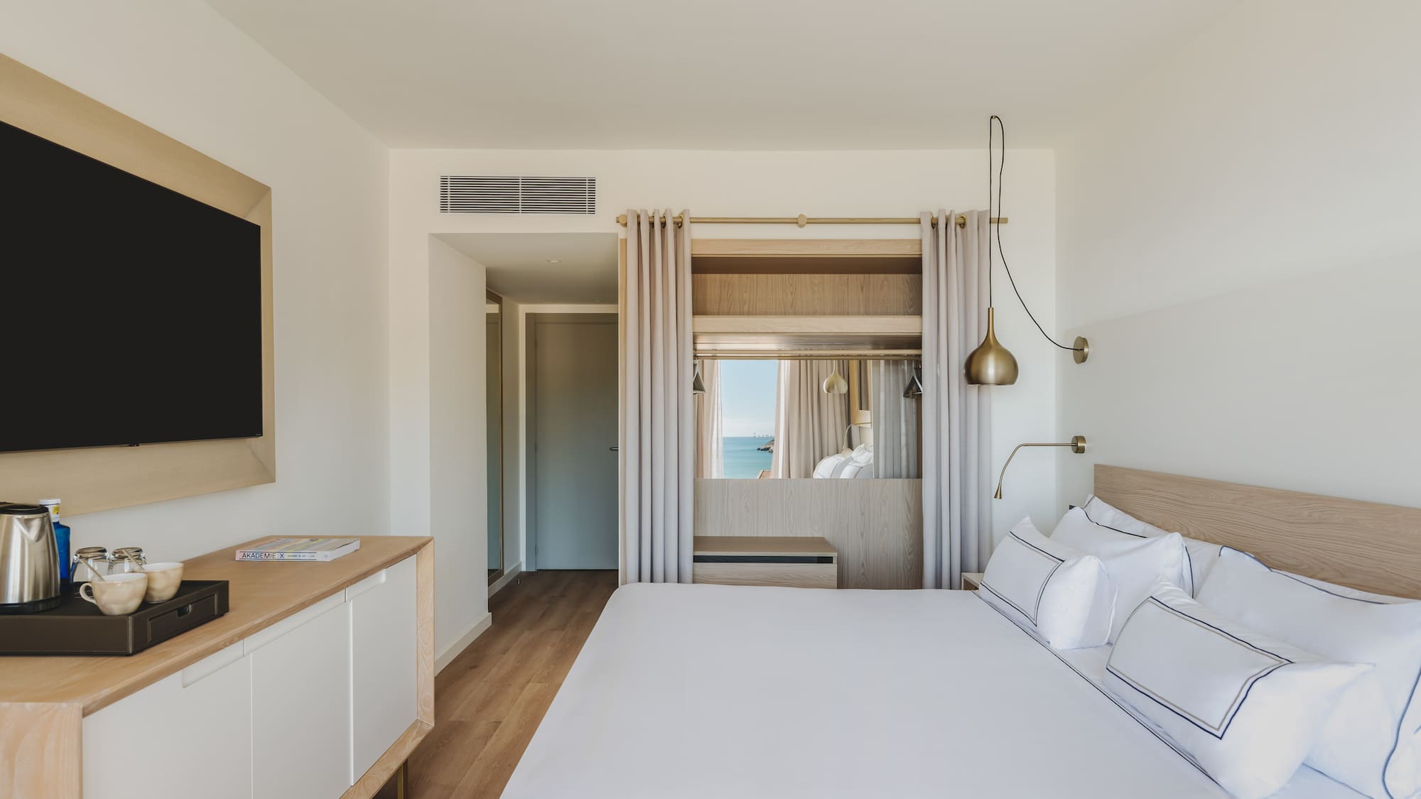 Modern hotel room with ocean view, white bed, wooden furniture, and sleek TV.