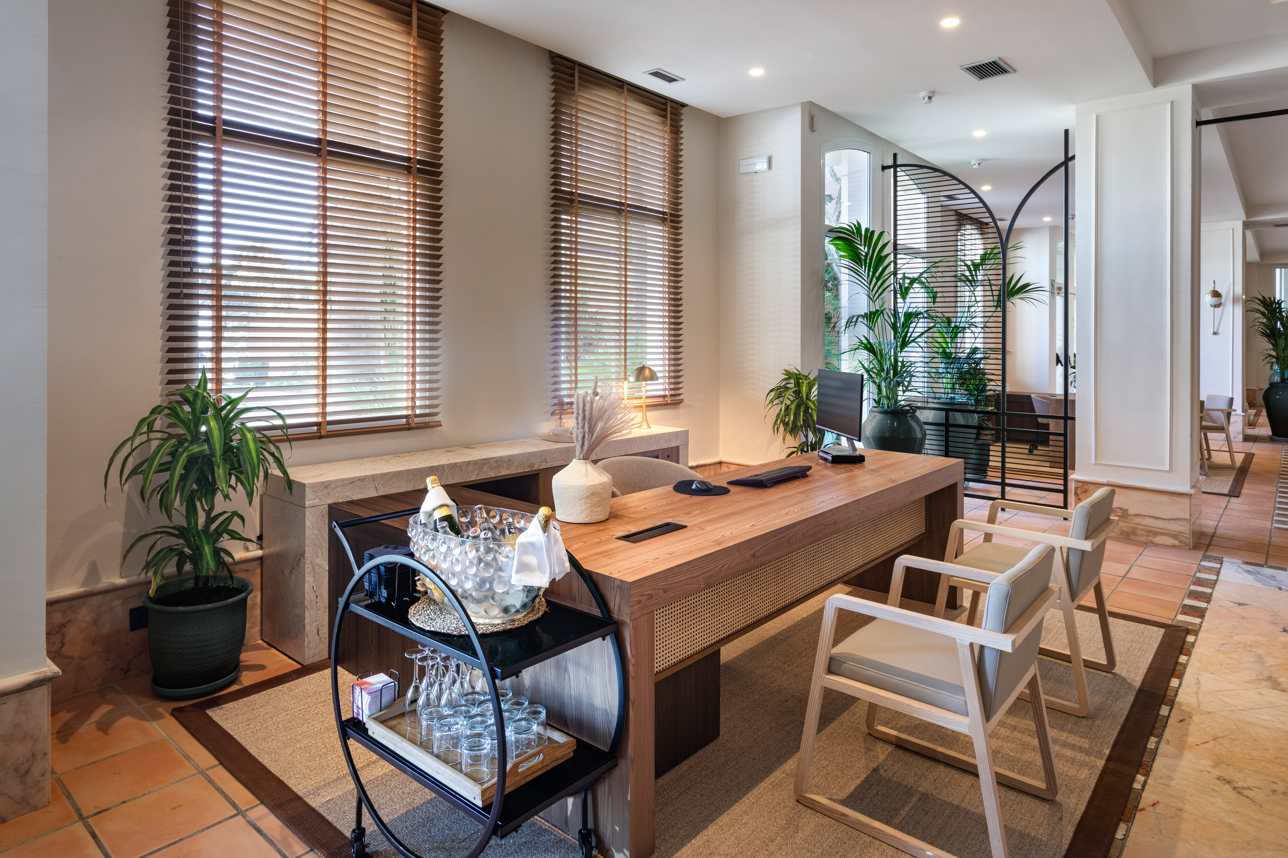 Modern hotel reception desk with wooden accents, bar cart, and indoor plants.