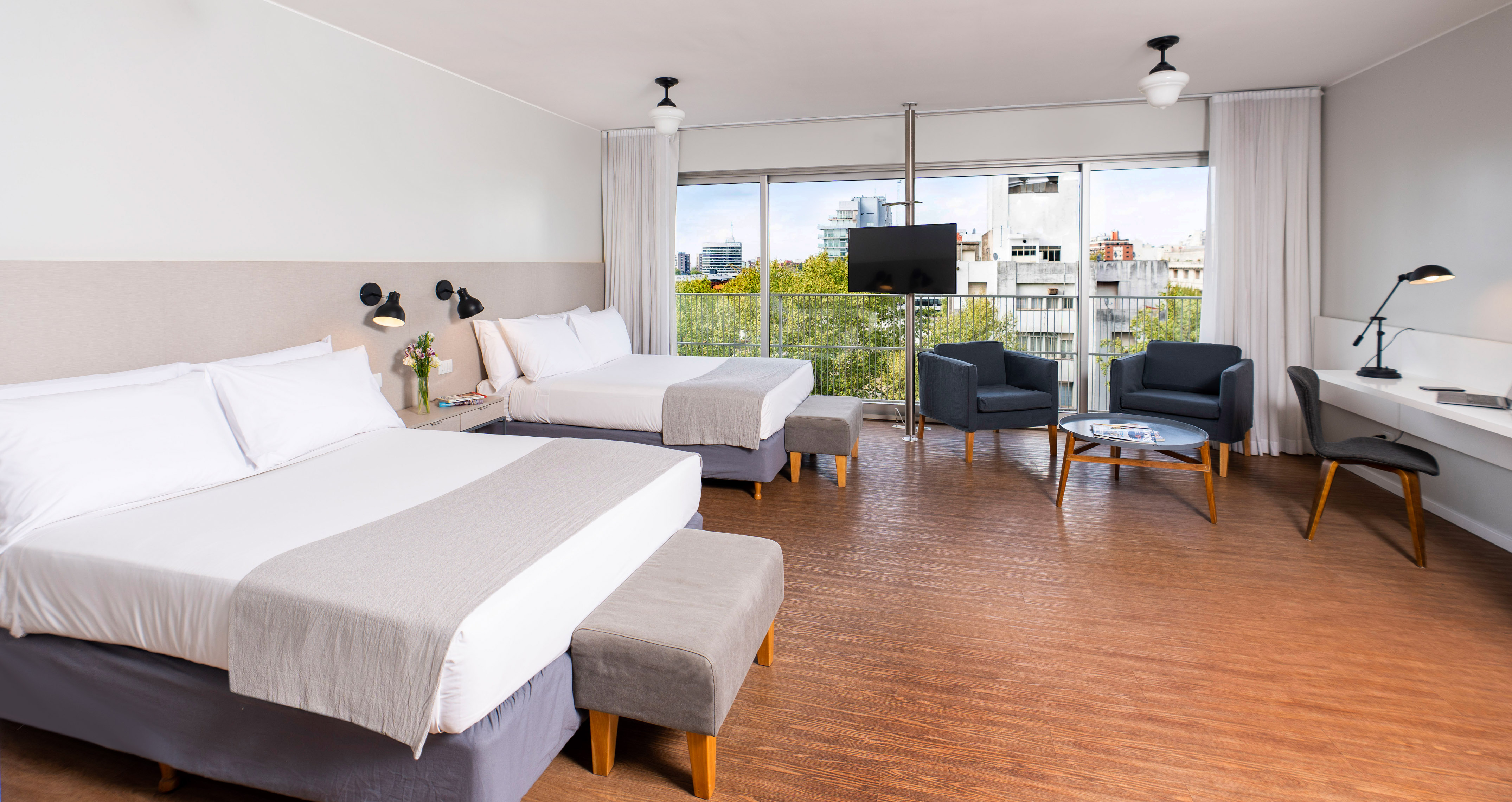 Modern hotel room with two beds, city view balcony, seating area, and desk.