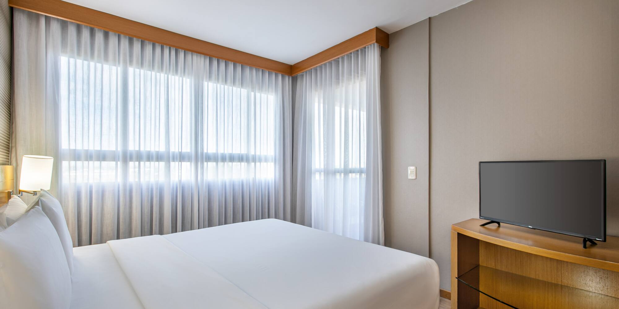 Modern hotel bedroom with large bed, sheer curtains, and flat-screen TV.