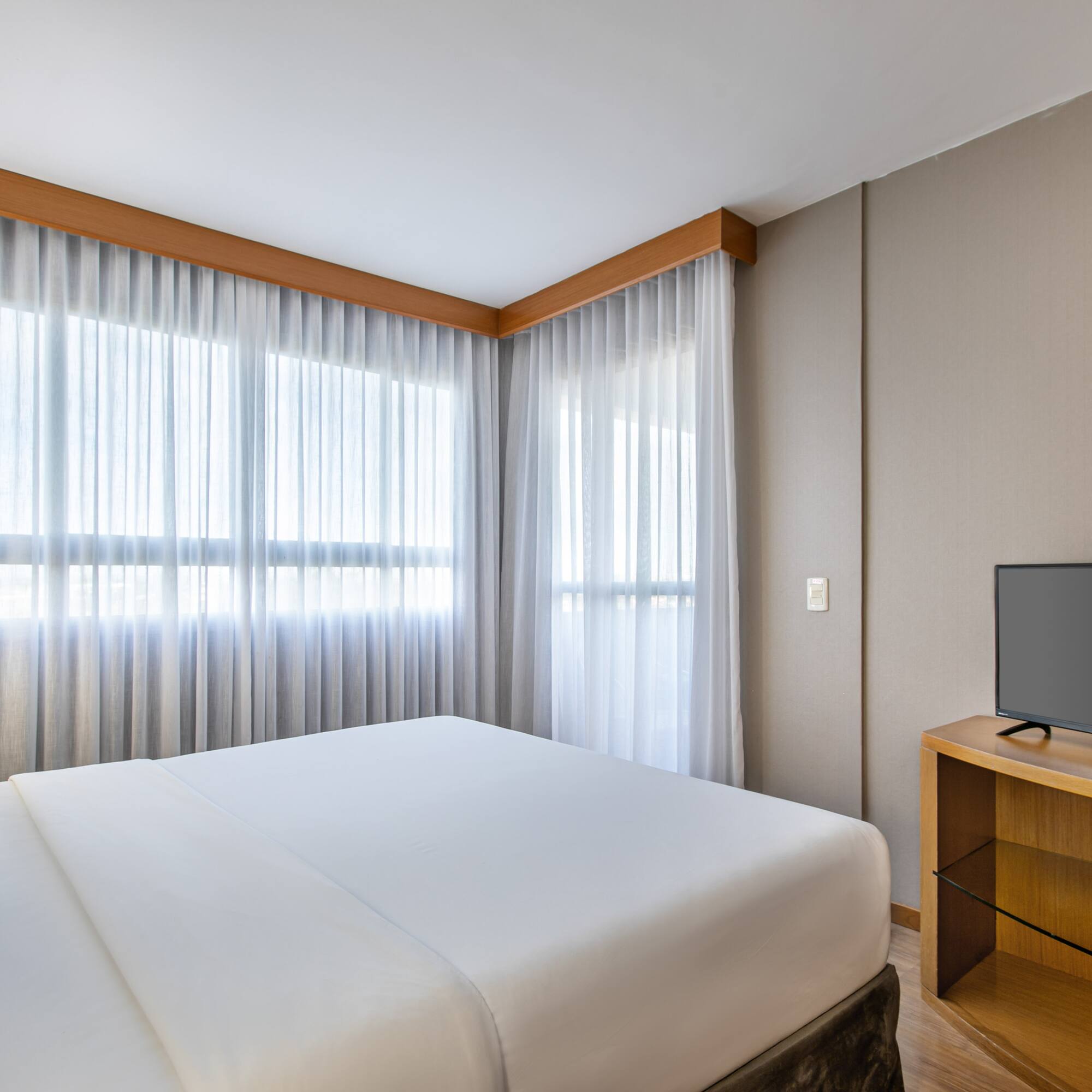 Modern hotel bedroom with large bed, sheer curtains, and flat-screen TV.