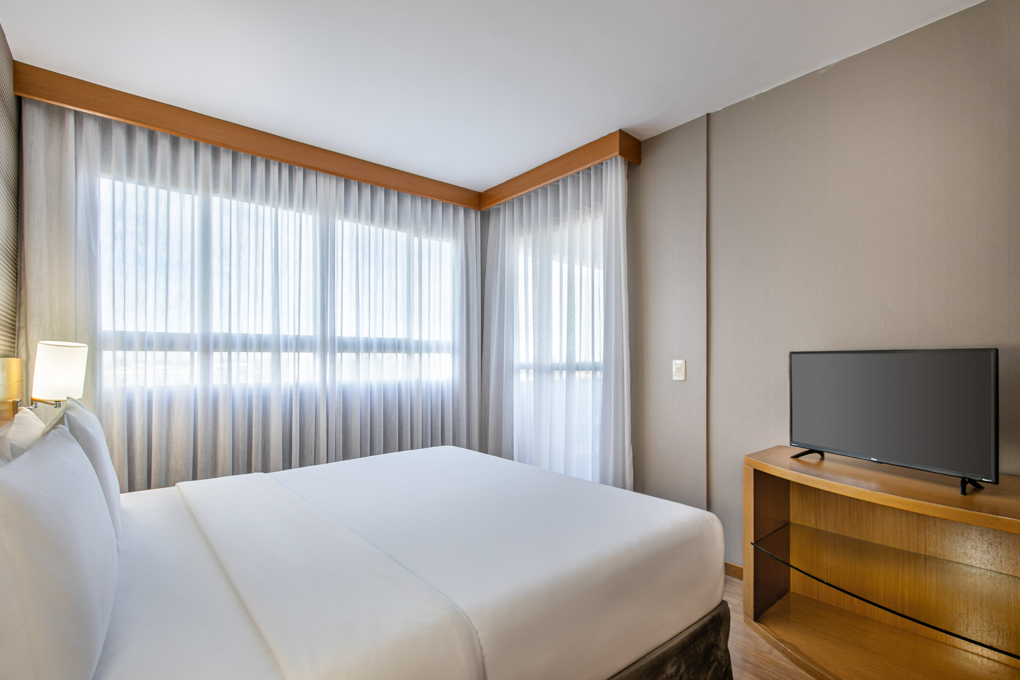 Modern hotel bedroom with large bed, sheer curtains, and flat-screen TV.