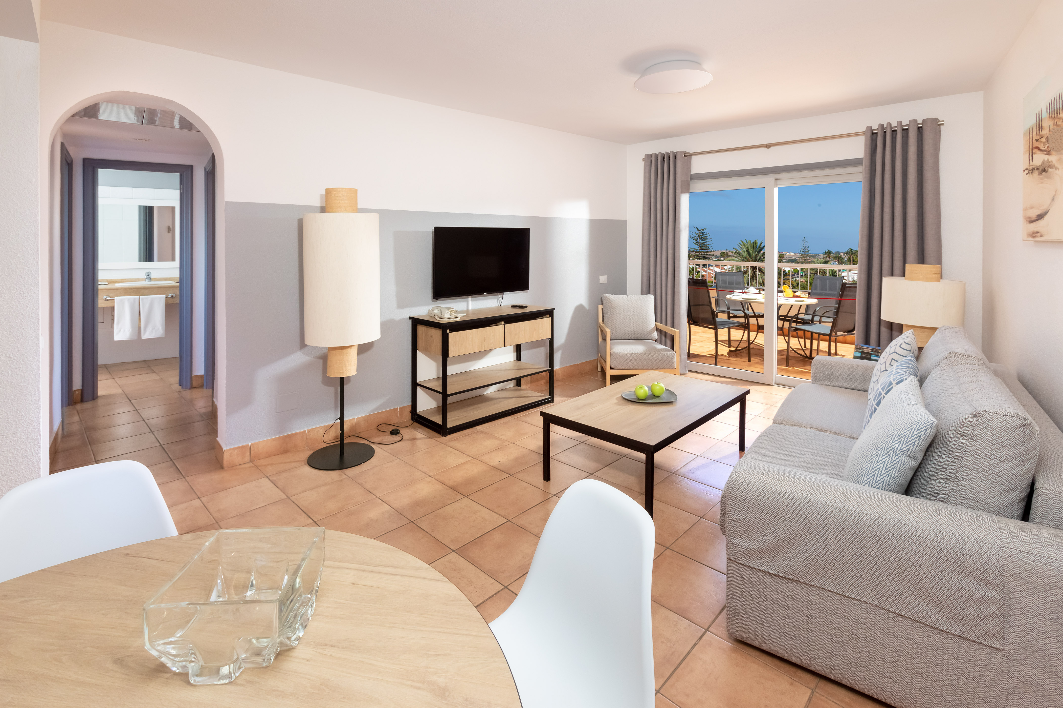 Bright living room with sofa, TV, dining table, and balcony overlooking palms.
