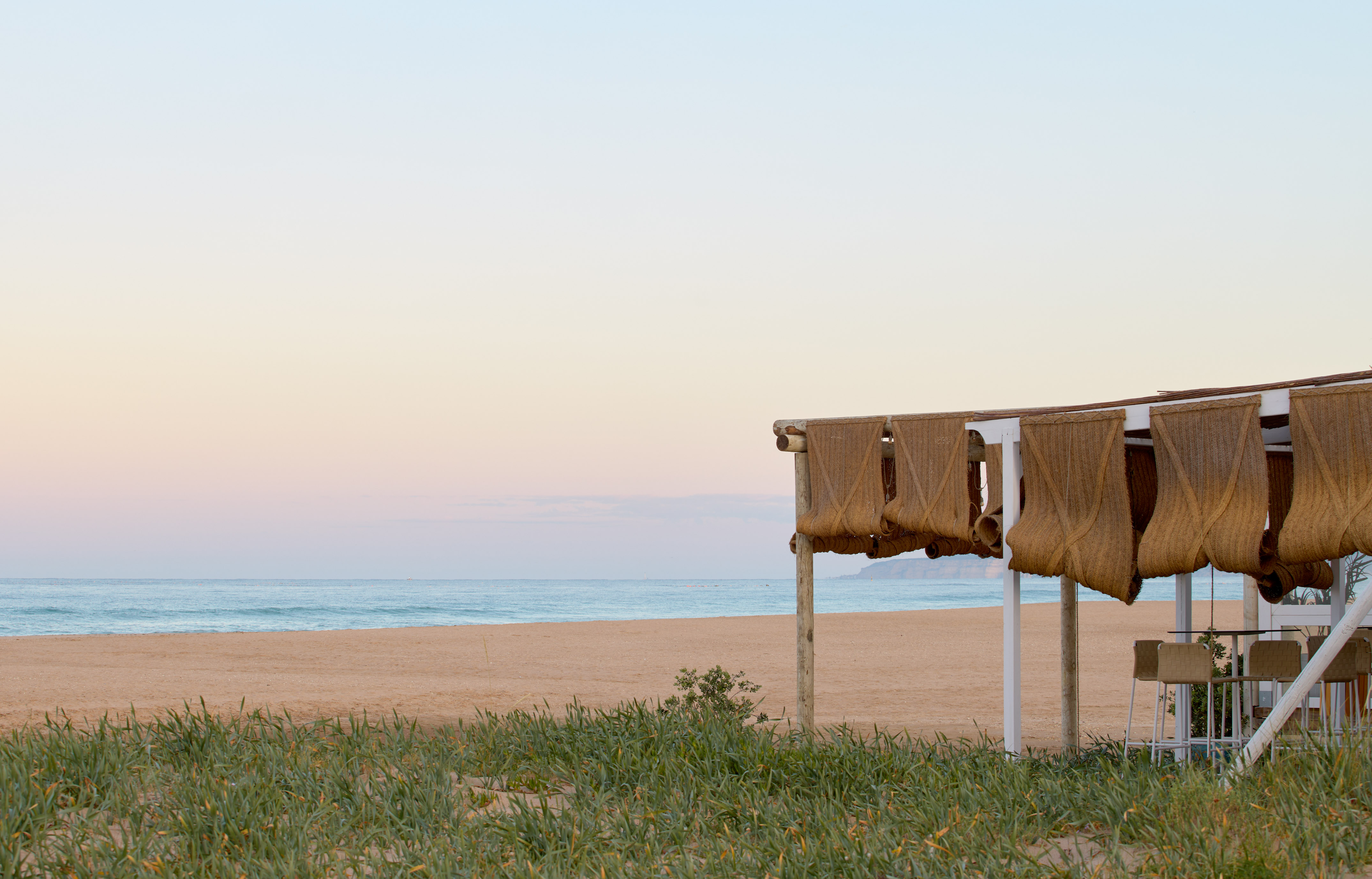 Beach bar with woven shades on sandy coast, calm ocean, pastel sky.