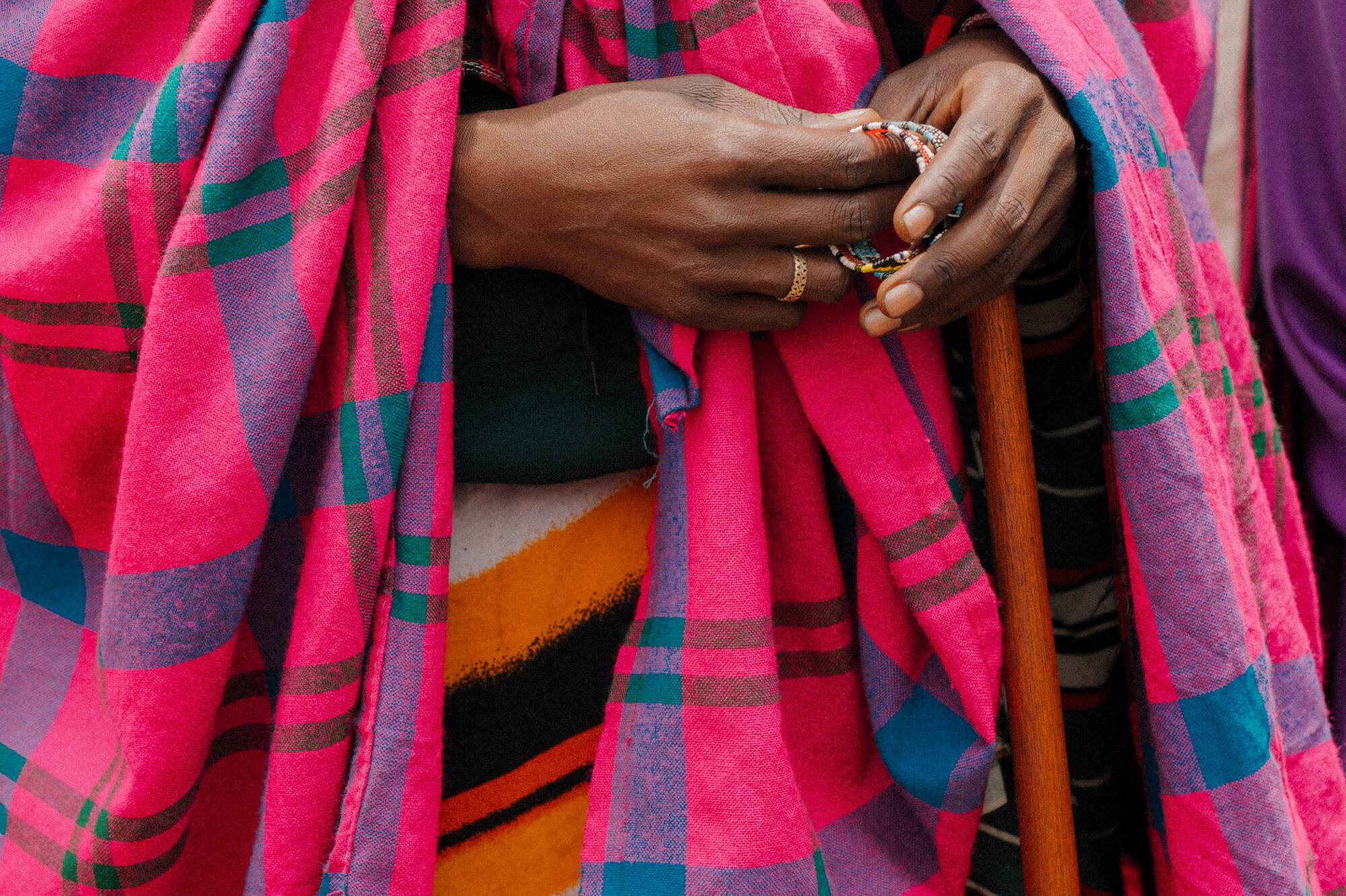 Hands holding beads and staff, draped in vibrant pink Maasai plaid shuka.