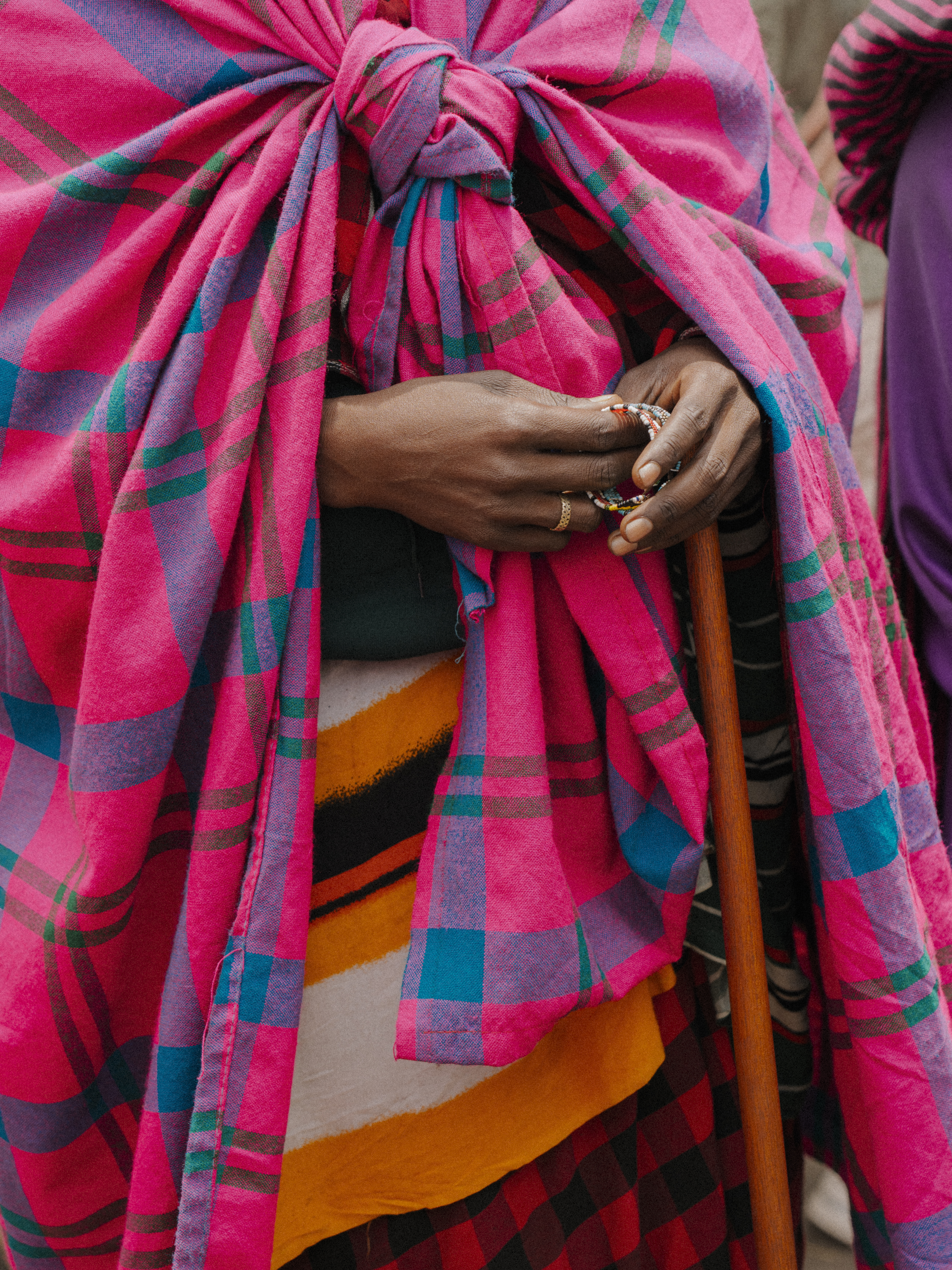 Hands holding beads and staff, draped in vibrant pink Maasai plaid shuka.