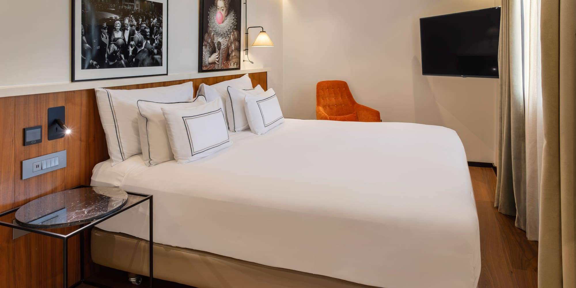 Modern hotel room with bed, diverse artworks, orange chair, and wall TV.