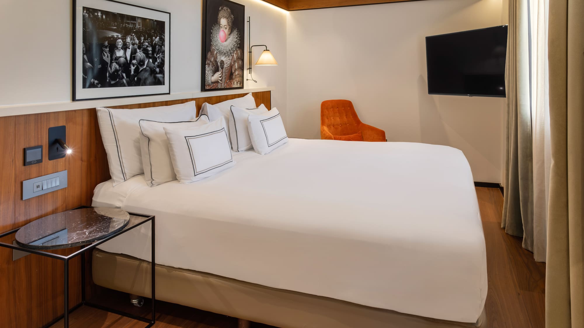 Modern hotel room with bed, diverse artworks, orange chair, and wall TV.