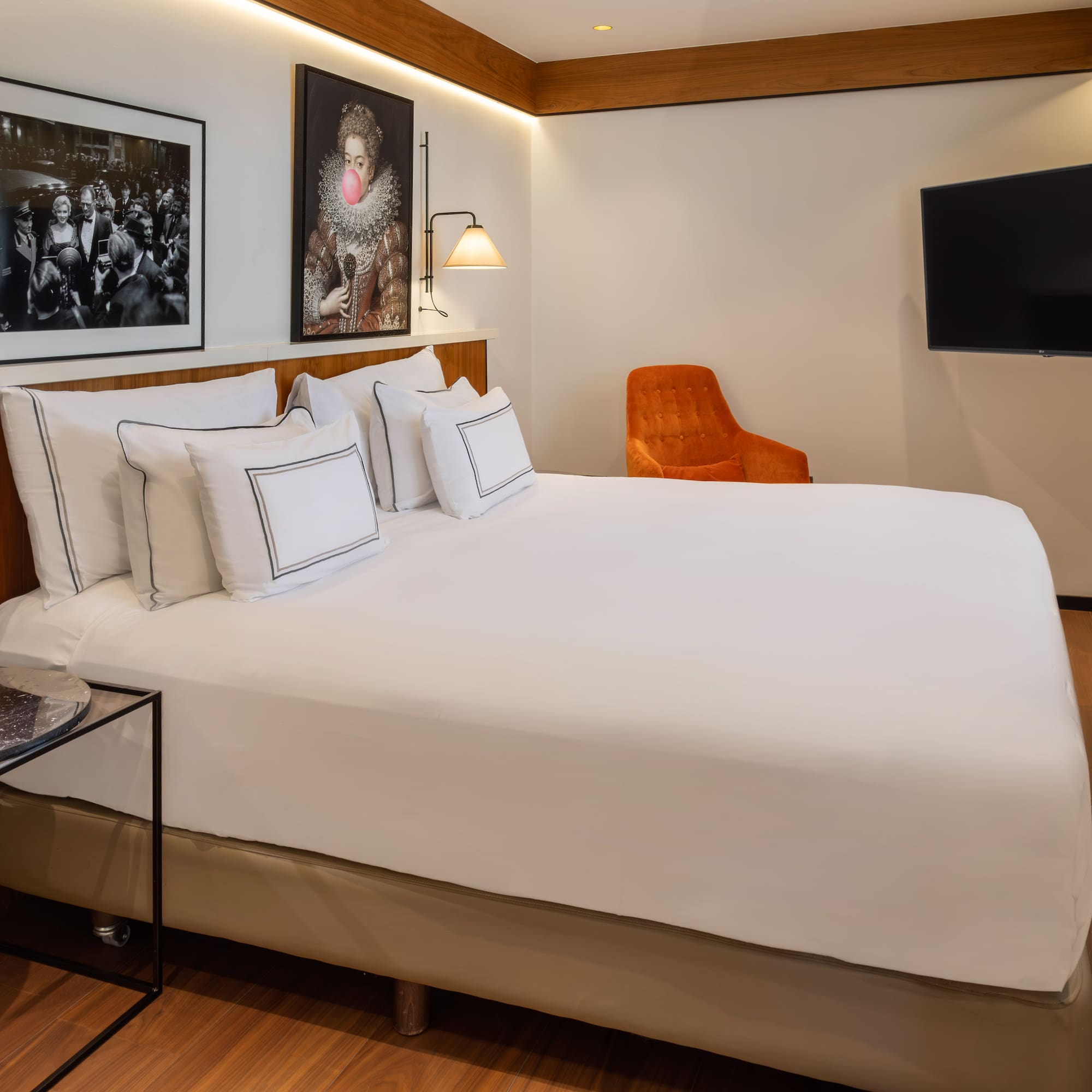 Modern hotel room with bed, diverse artworks, orange chair, and wall TV.