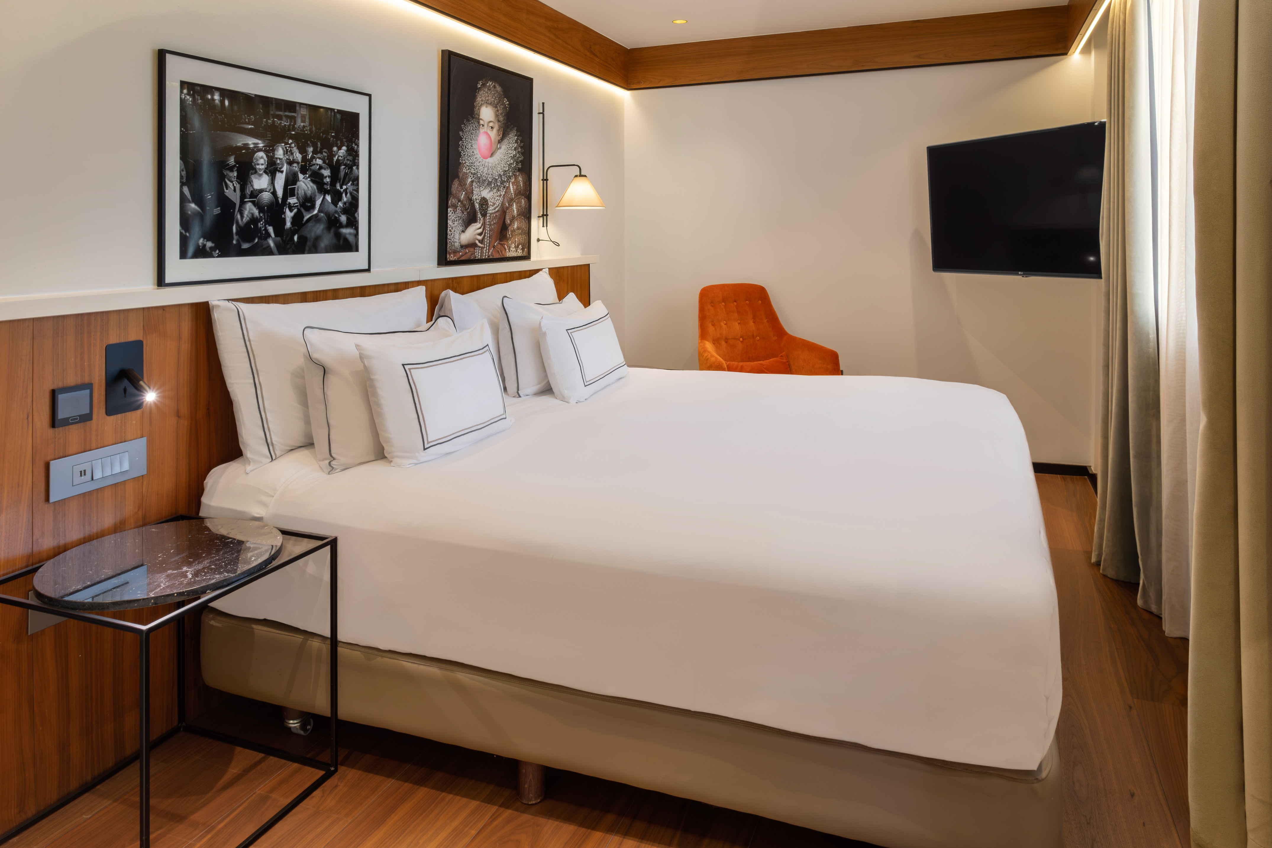 Modern hotel room with bed, diverse artworks, orange chair, and wall TV.