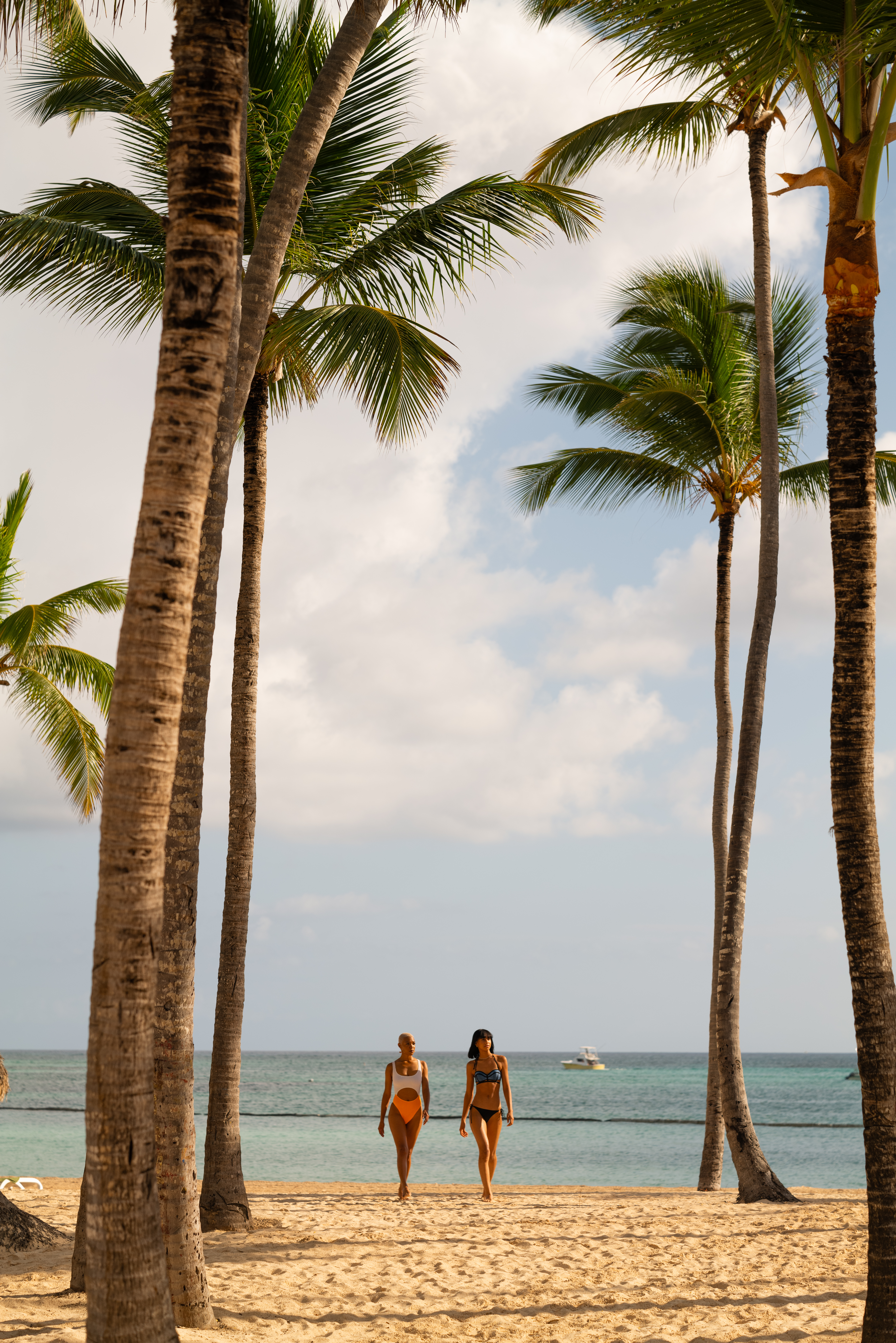 Two women in swimsuits stroll on a sunny tropical beach with palm trees.