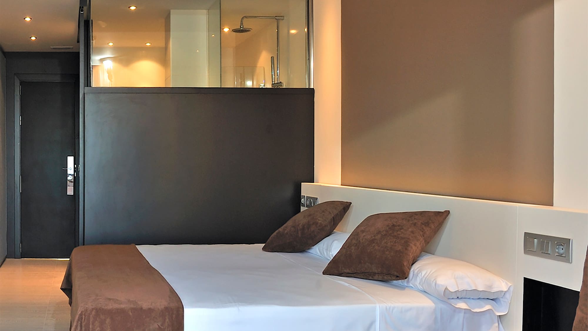 Modern hotel room with brown and white bed, sofa, and glass shower.