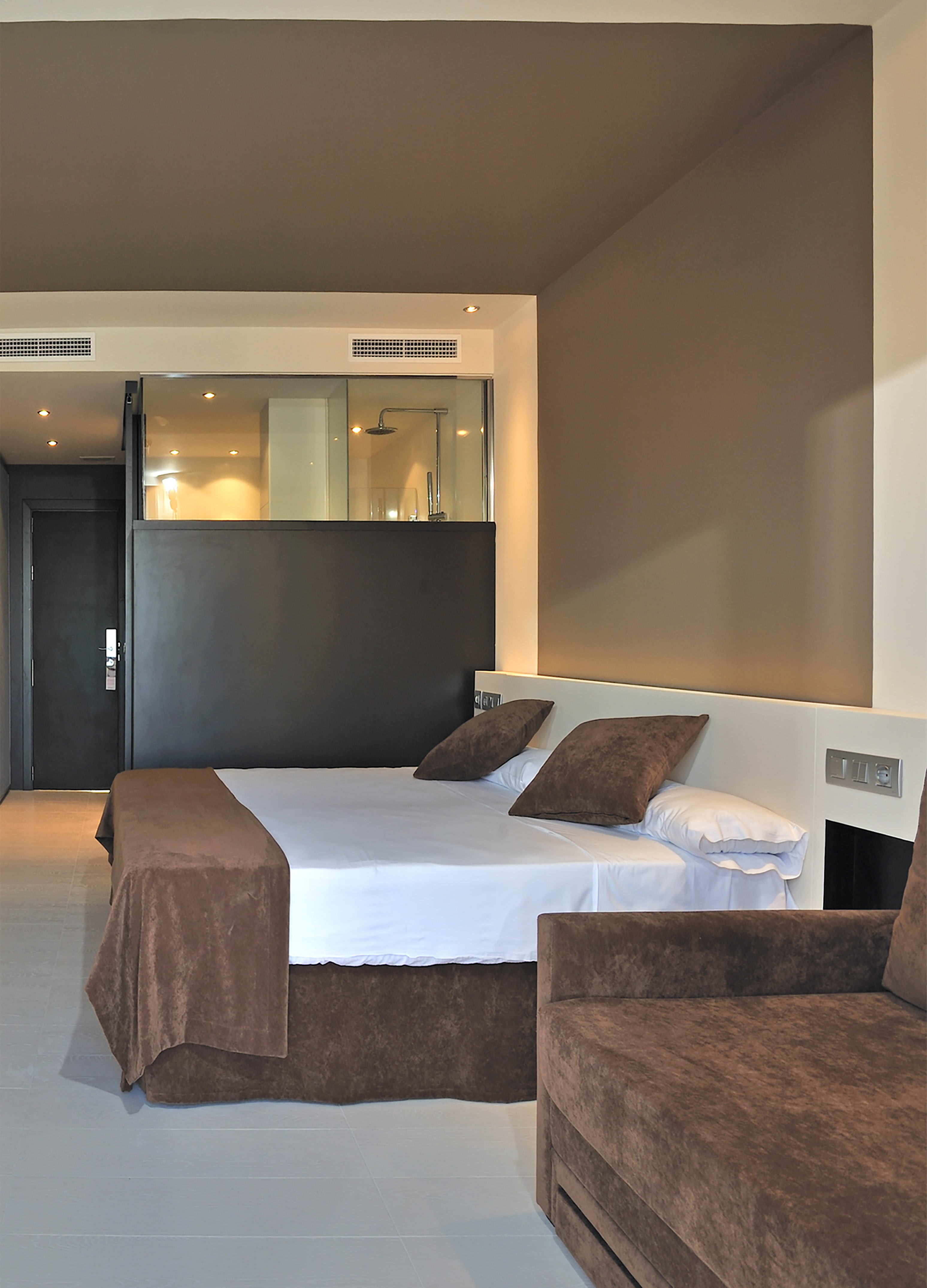 Modern hotel room with brown and white bed, sofa, and glass shower.