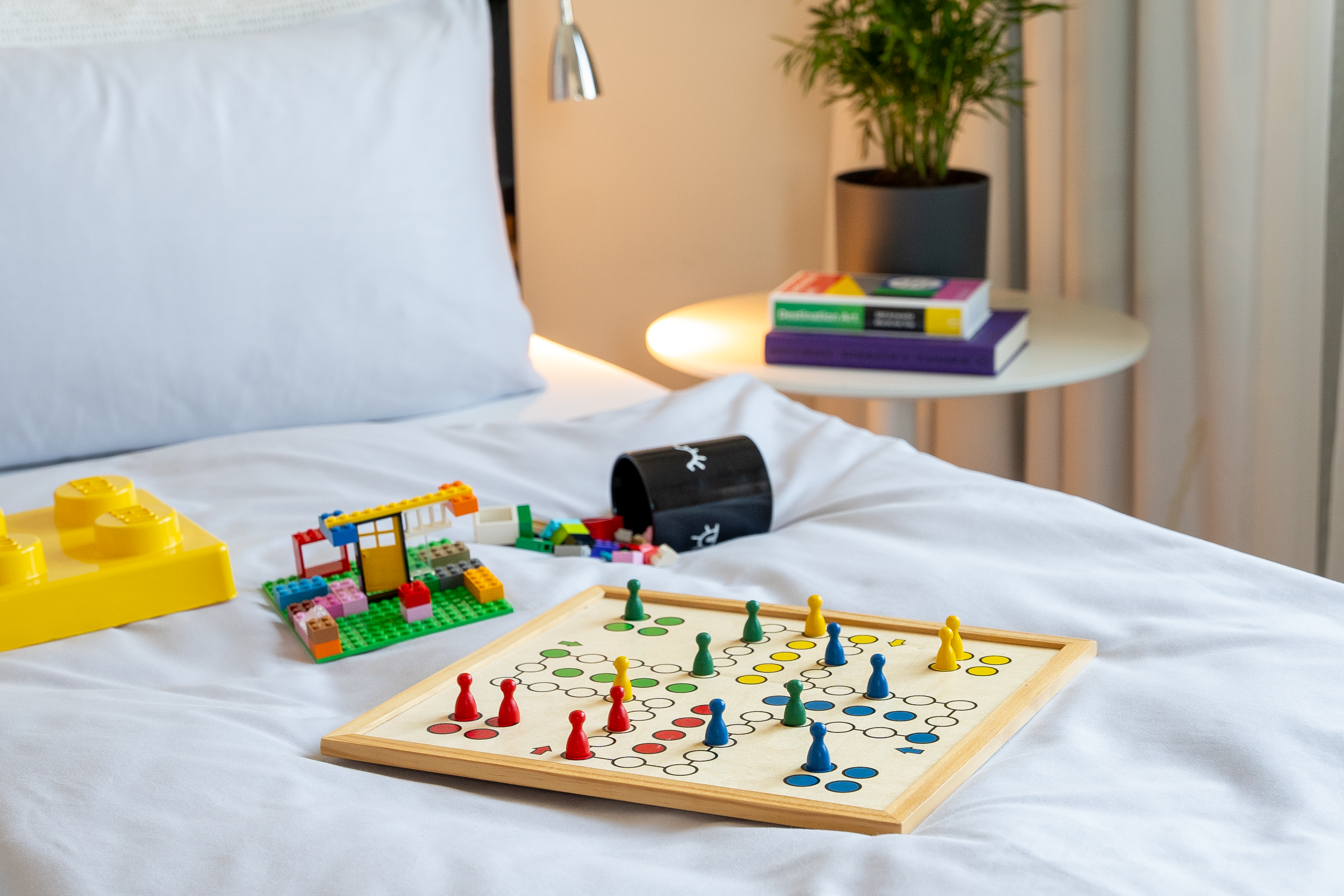 Ludo board game, colorful Lego bricks, and toys on a white hotel bed.