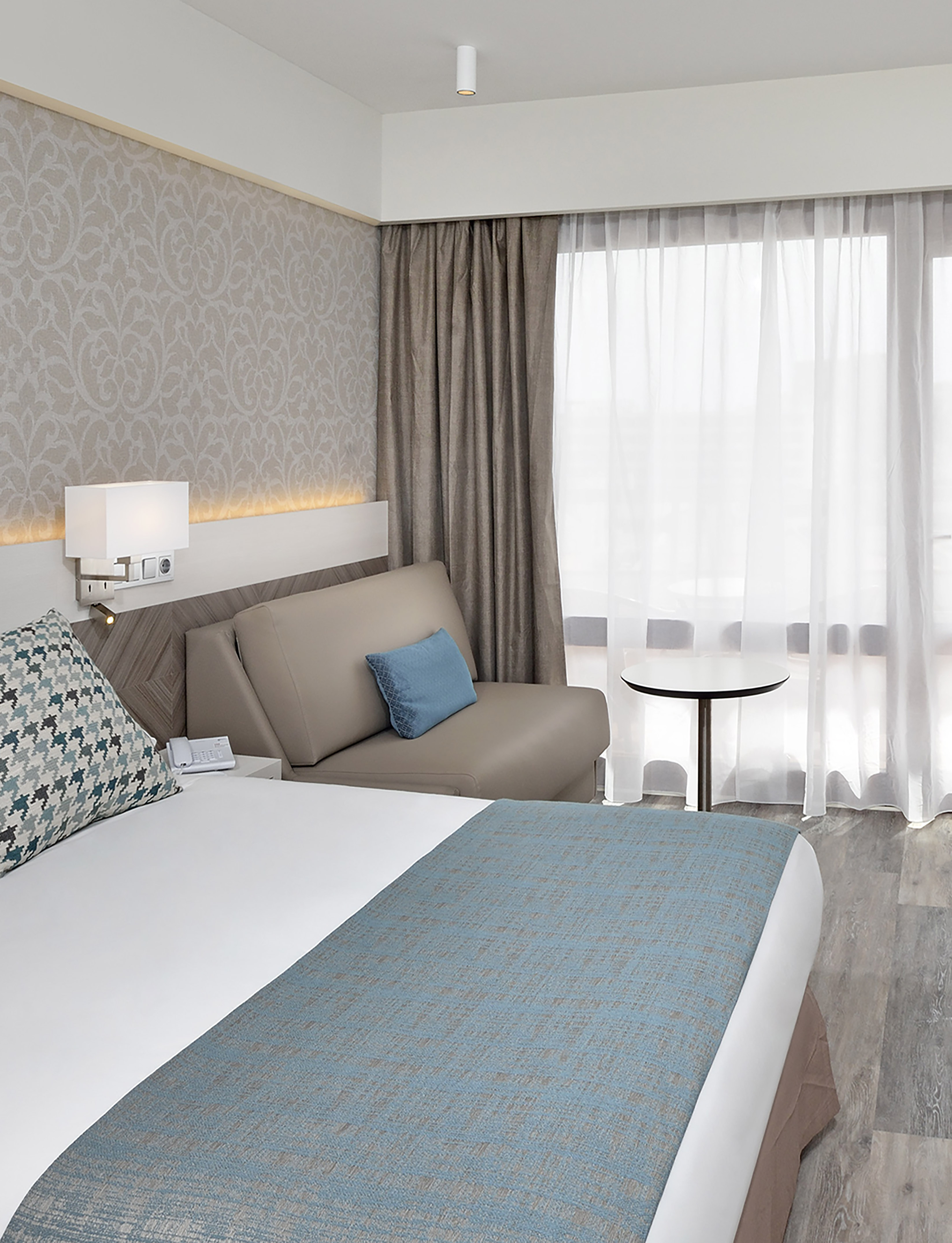 Modern hotel room with bed, cozy sofa, patterned wallpaper, and bright window.