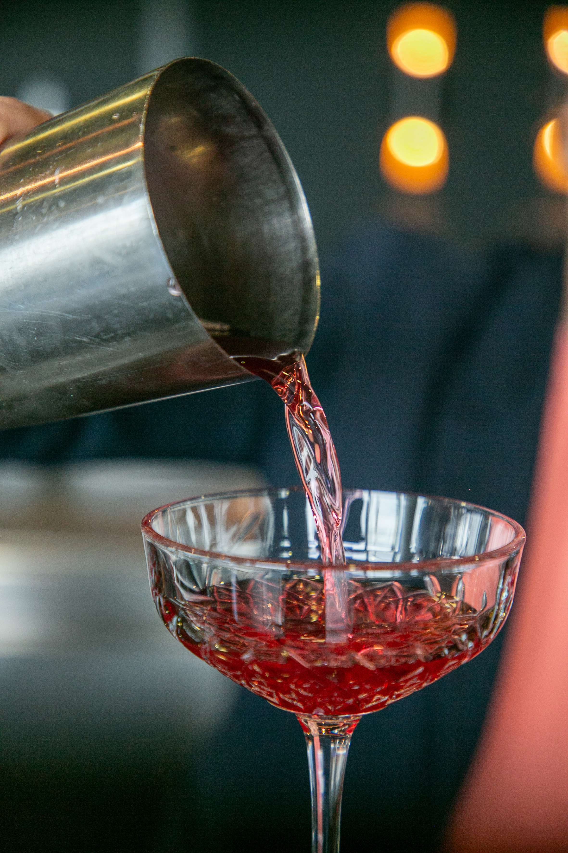 Bartender pouring red cocktail from shaker into stemmed glass.
