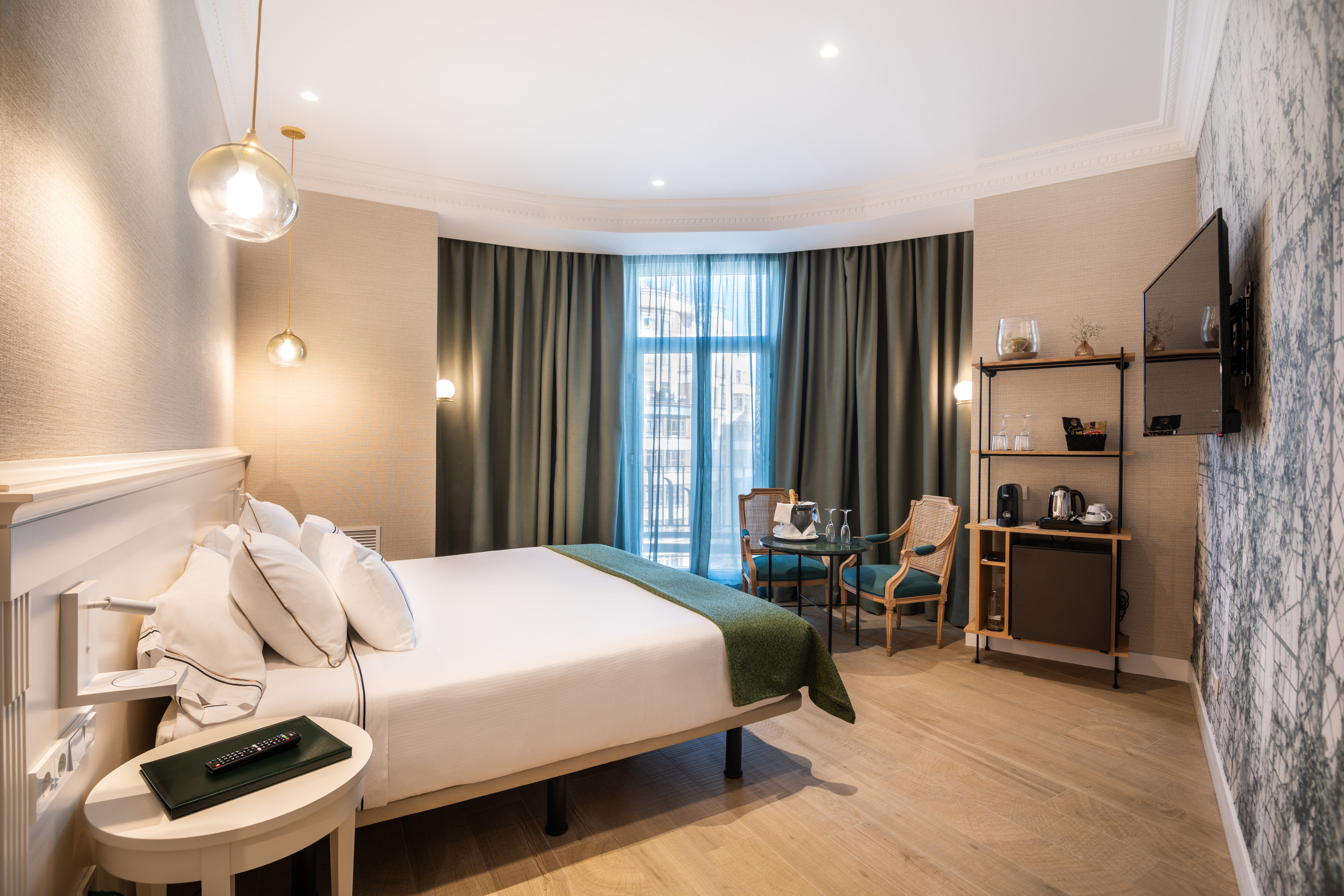 Modern hotel room with a large bed, bay window, minibar, and seating area.