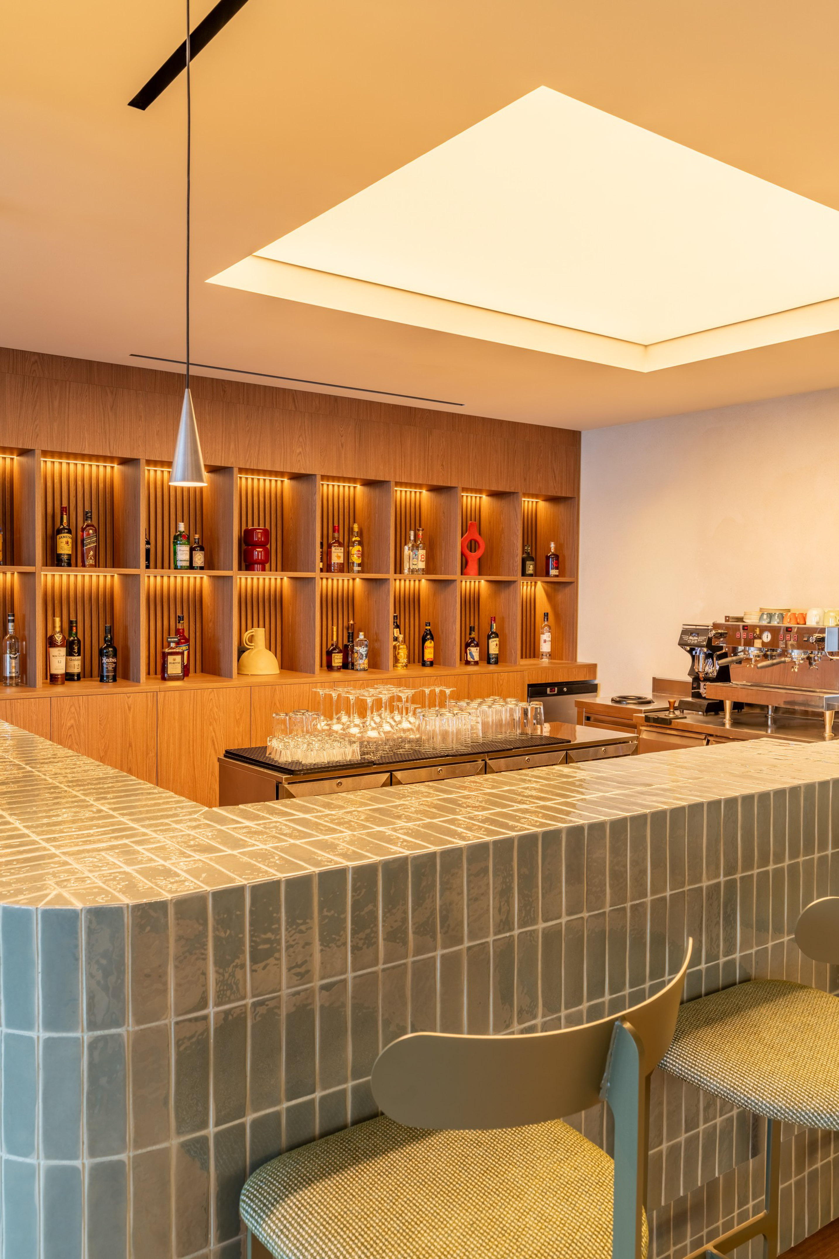 Modern bar with green tiled counter, wooden liquor shelves, and espresso machine.