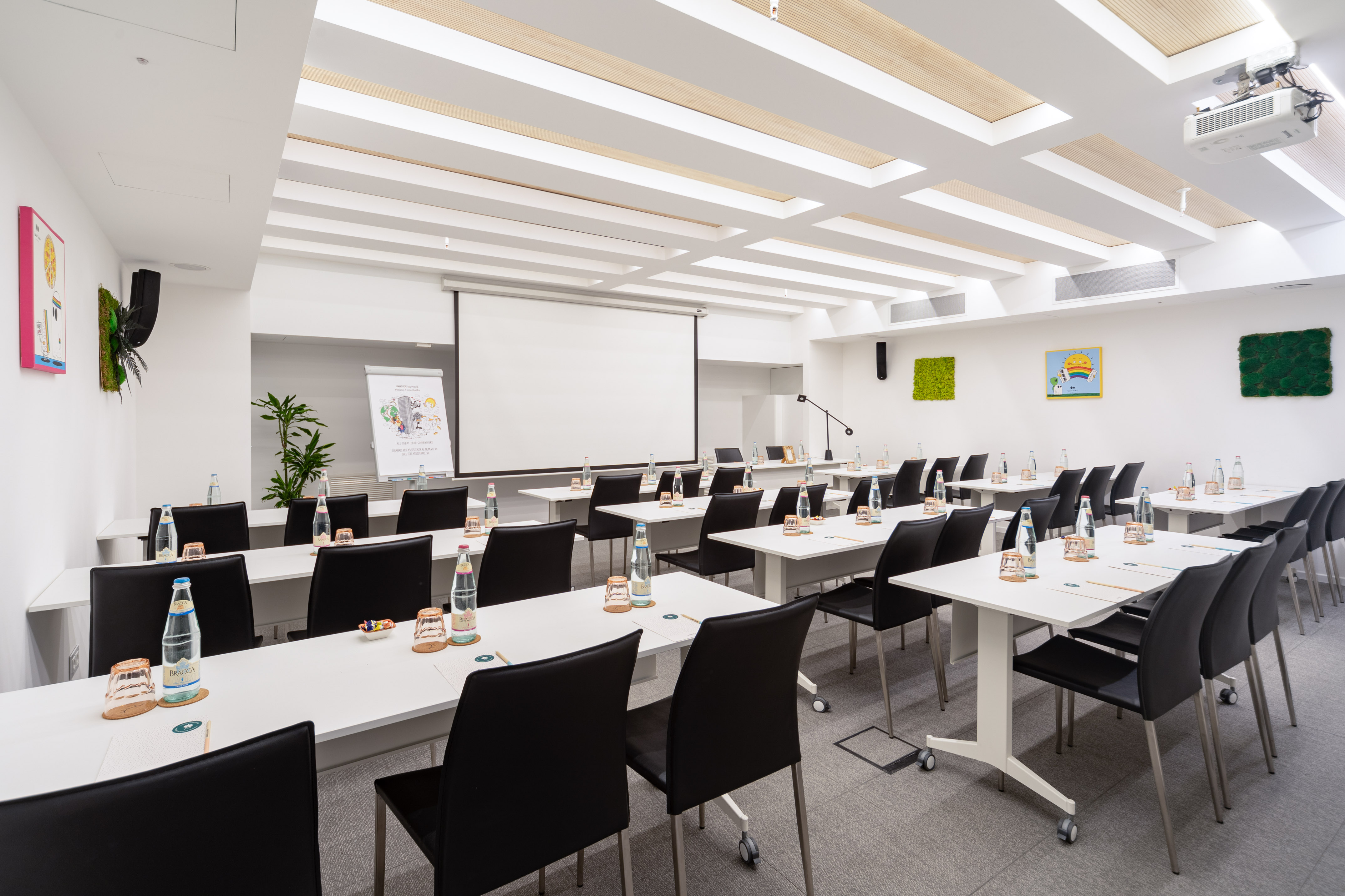 Modern bright meeting room with projector screen, tables, and black chairs.