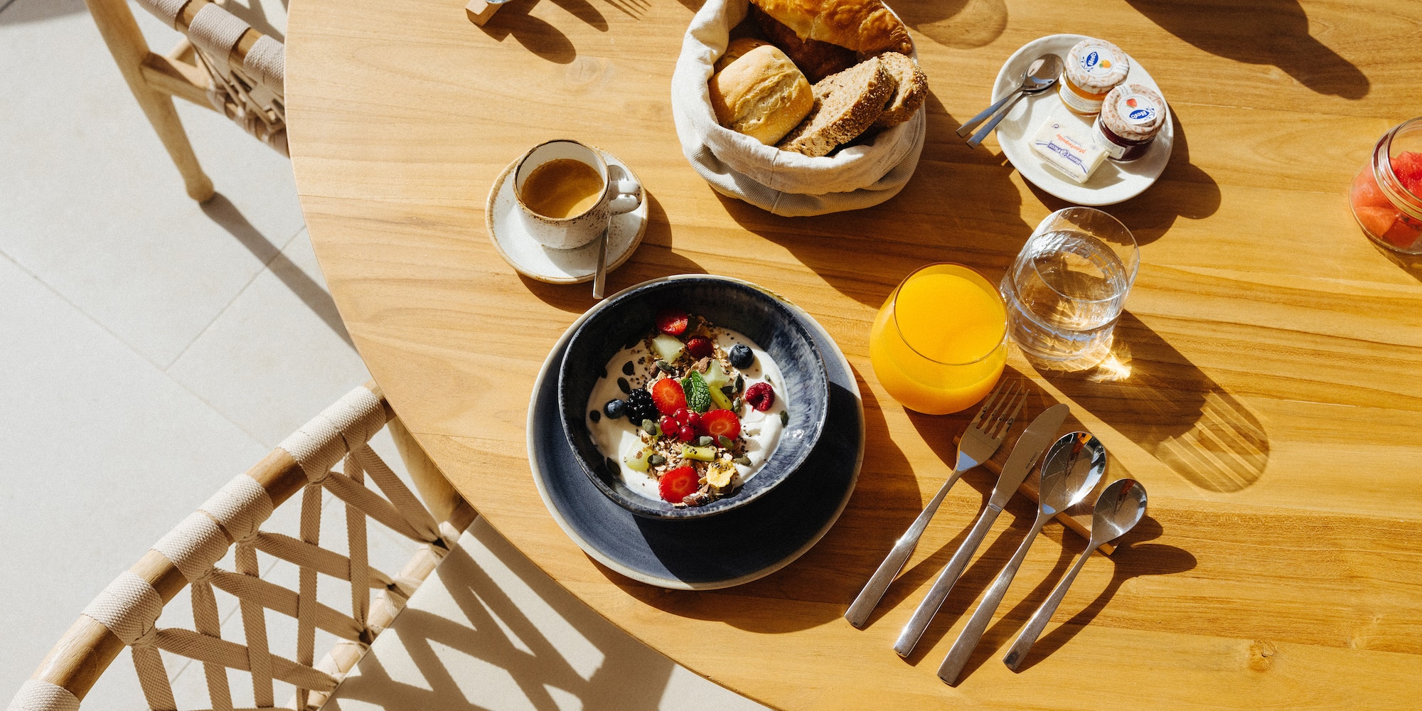 Sunny breakfast on wooden table with yogurt, berries, coffee, juice, bread.